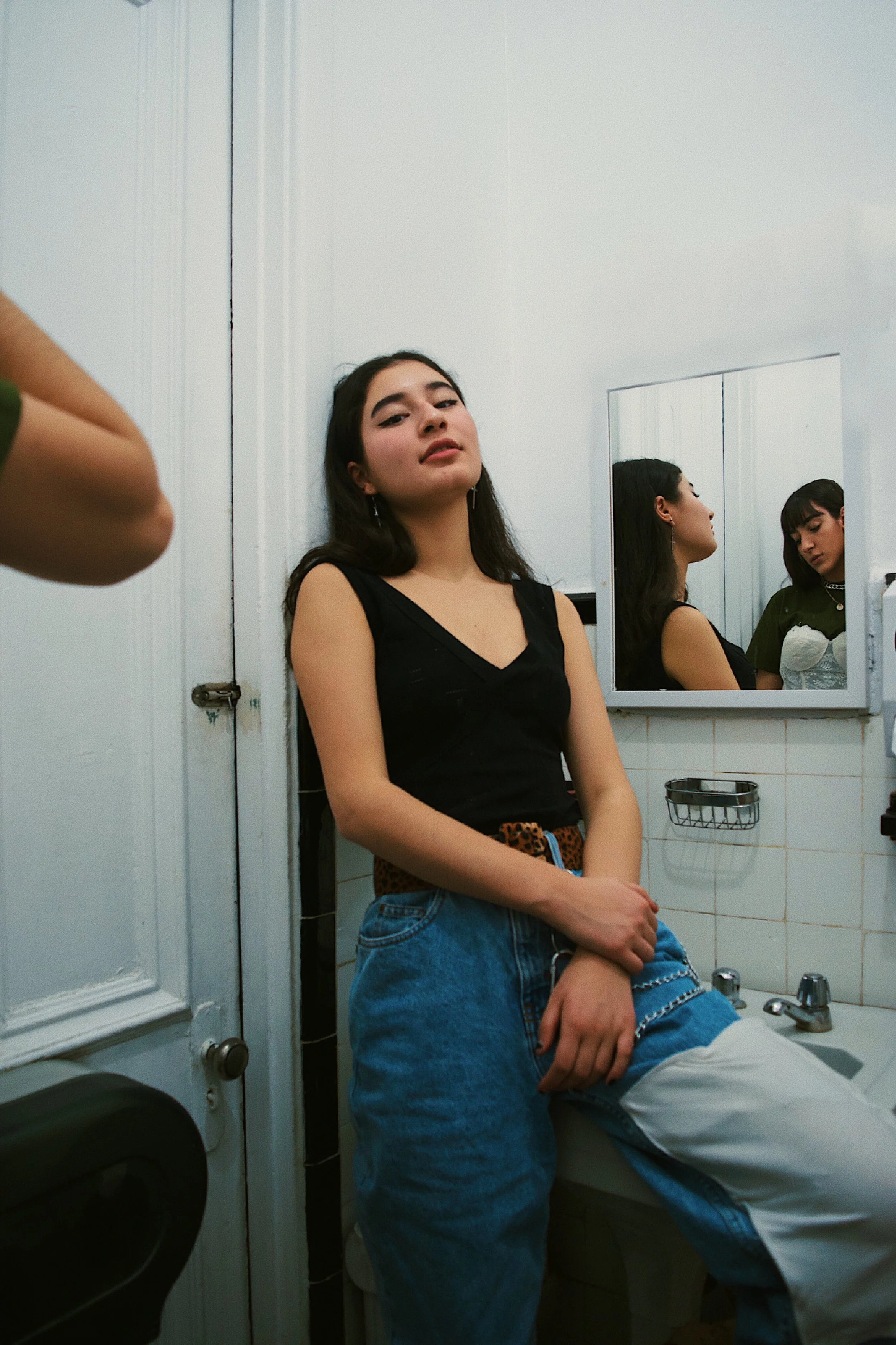 A young woman standing in a bathroom with her back against the wall, looking at the camera. There is a mirror behind her reflecting her face and another young woman in a white top. The bathroom has white walls, a sink, and a small shelf.