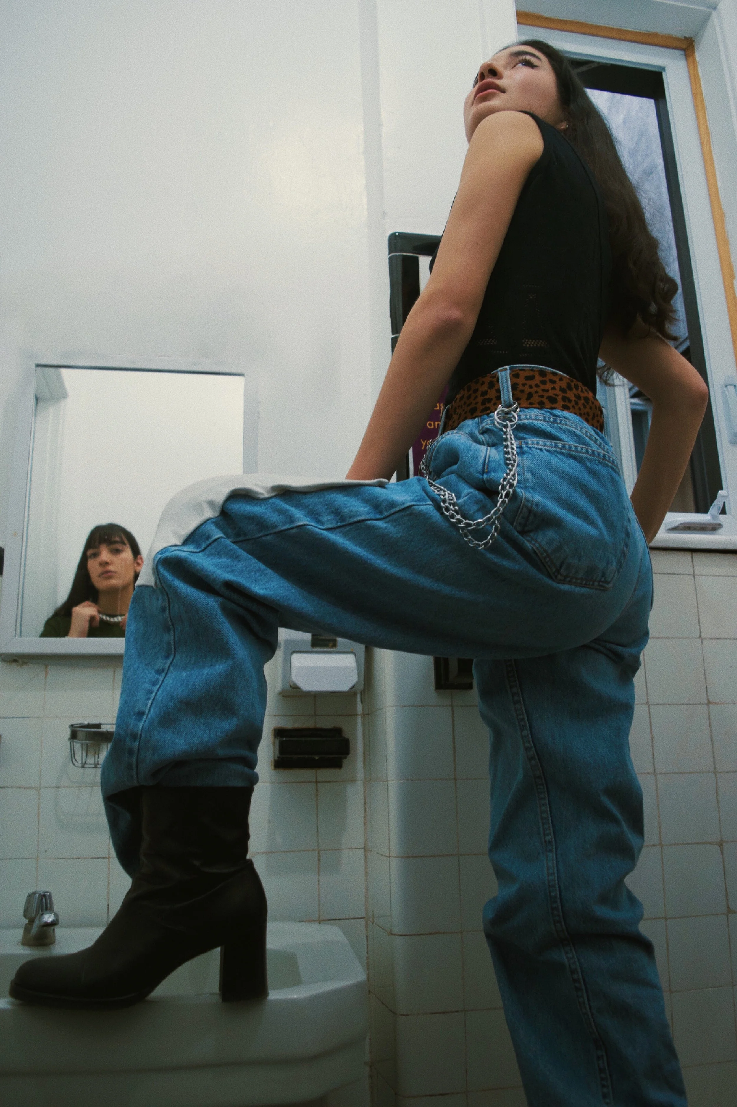 A young woman poses with one foot on a toilet, wearing black heeled boots, blue jeans, a black tank top, and a leopard print belt, in a bathroom with a mirror showing her reflection.