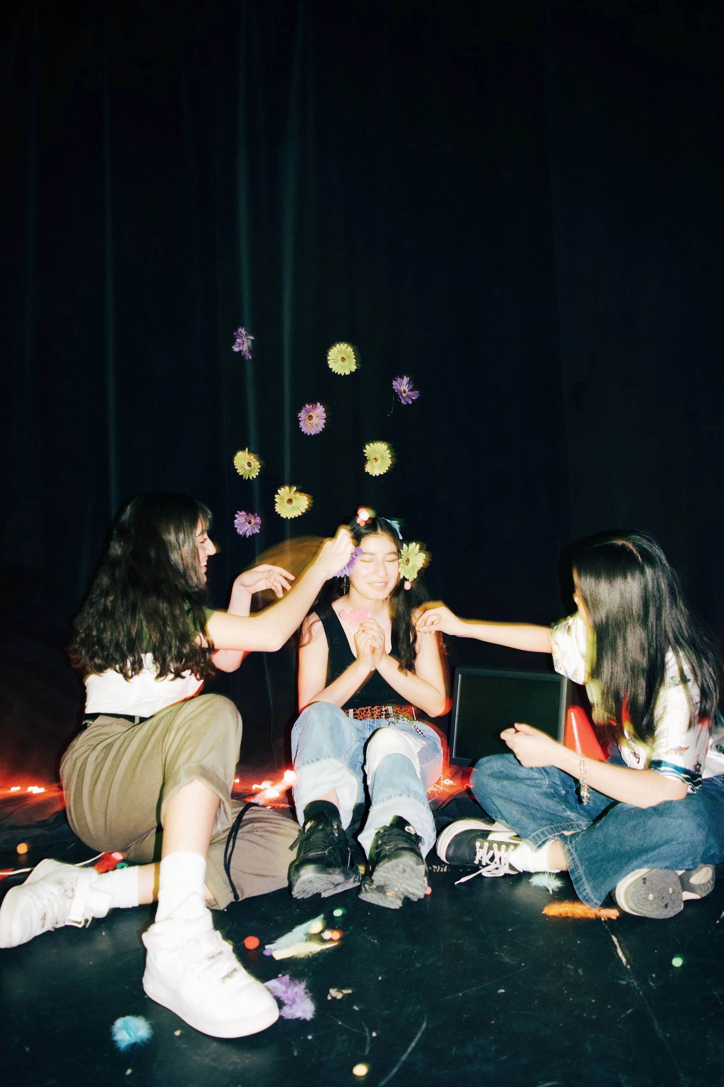 Three young girls sitting on the floor in front of a dark curtain, with falling flower petals and string lights, enjoying a playful moment.