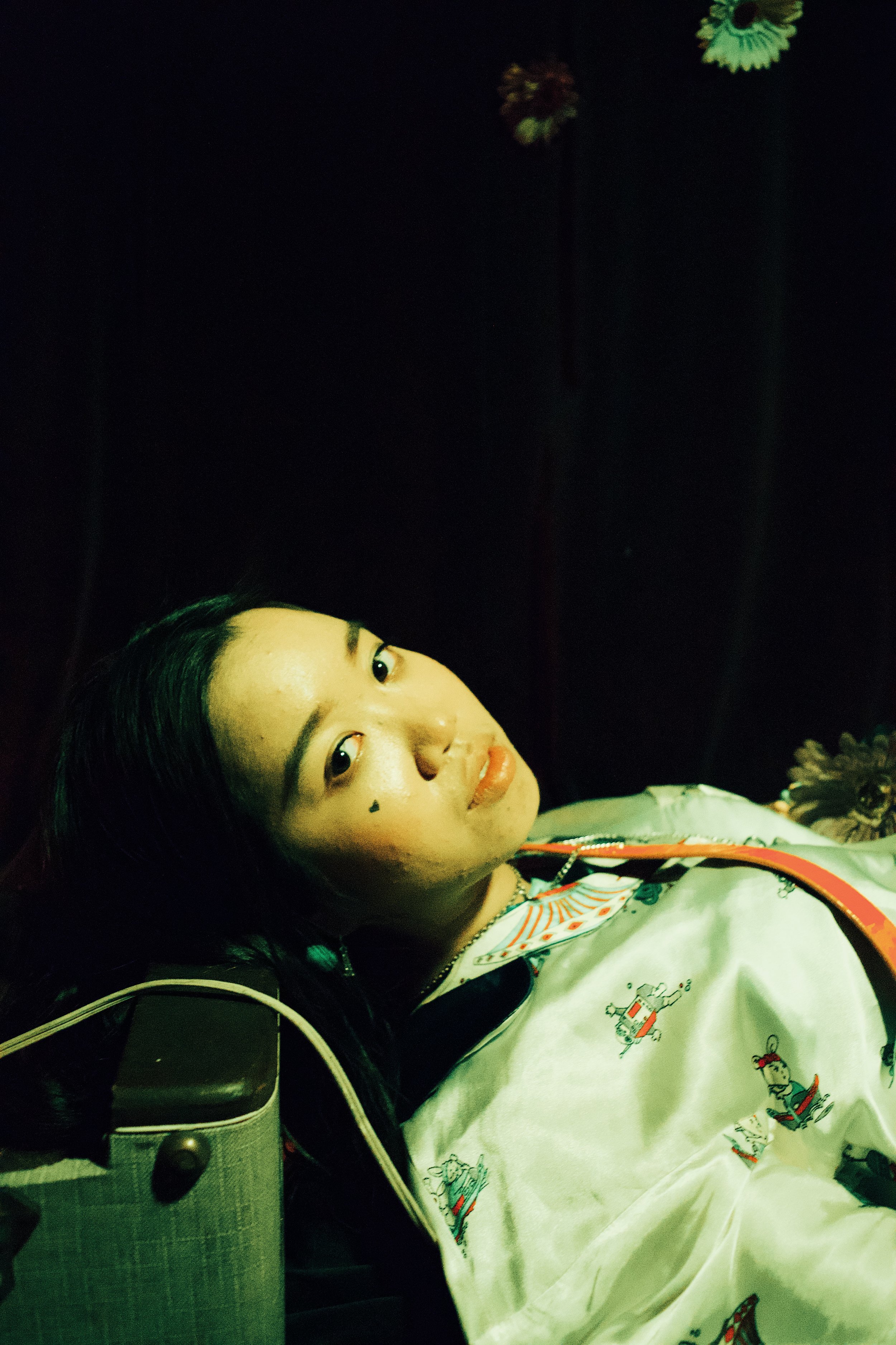 A woman lying down against a dark background, with long black hair and wearing a white garment with colorful embroidery.