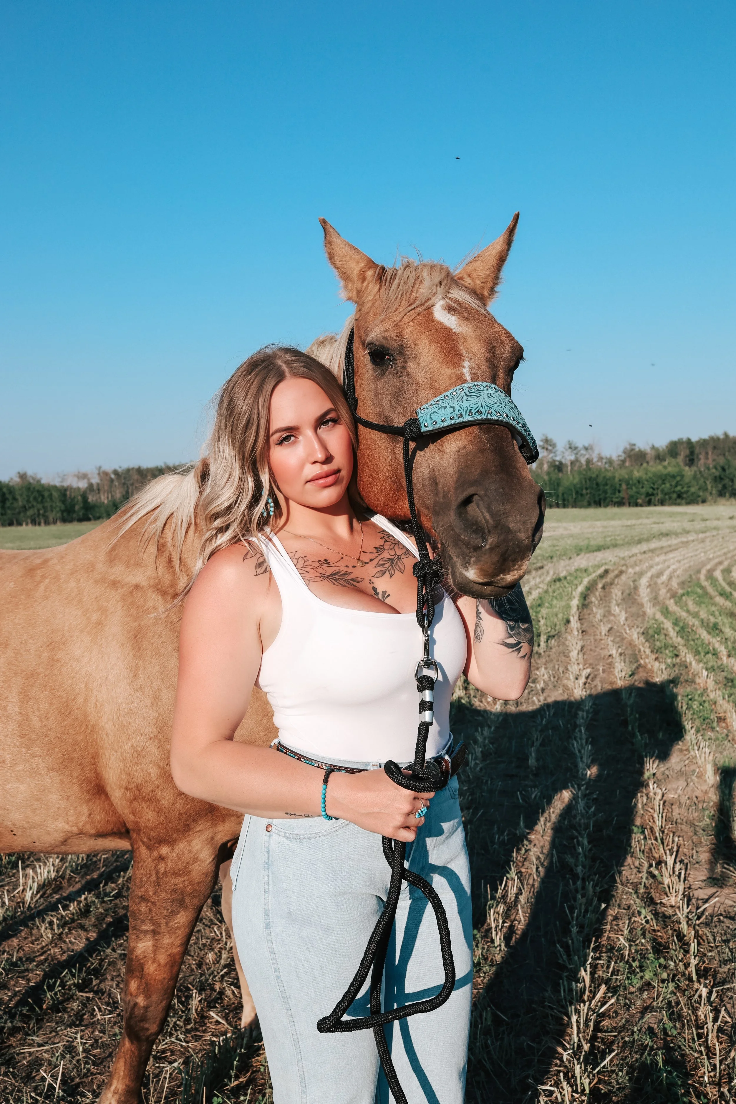 A young woman with blonde hair, tattoos, and a white tank top holding a black rope is standing next to a brown horse with a blue halter in a field on a sunny day.
