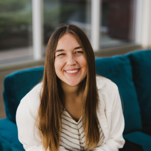 Smiling woman with long brown hair sitting on a blue couch in a well-lit room.