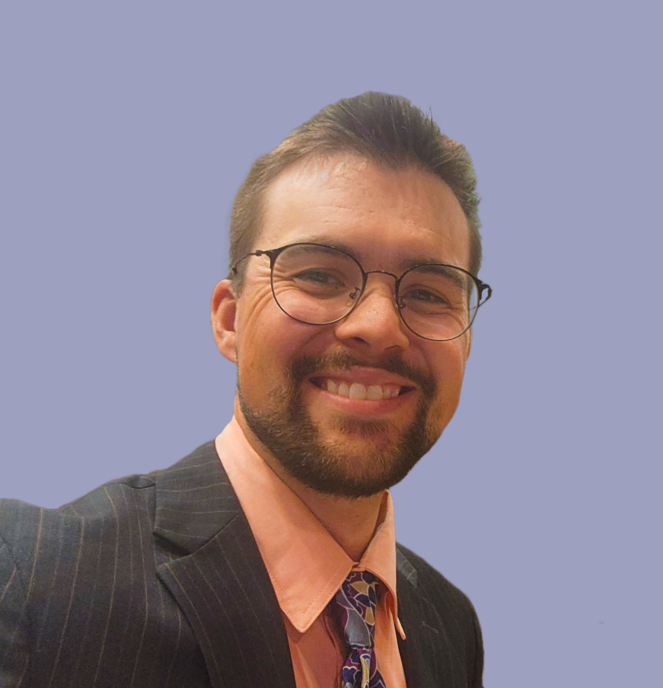 Close-up of a smiling man with glasses, wearing a dark pinstripe suit, orange shirt, and patterned tie, against a plain background.
