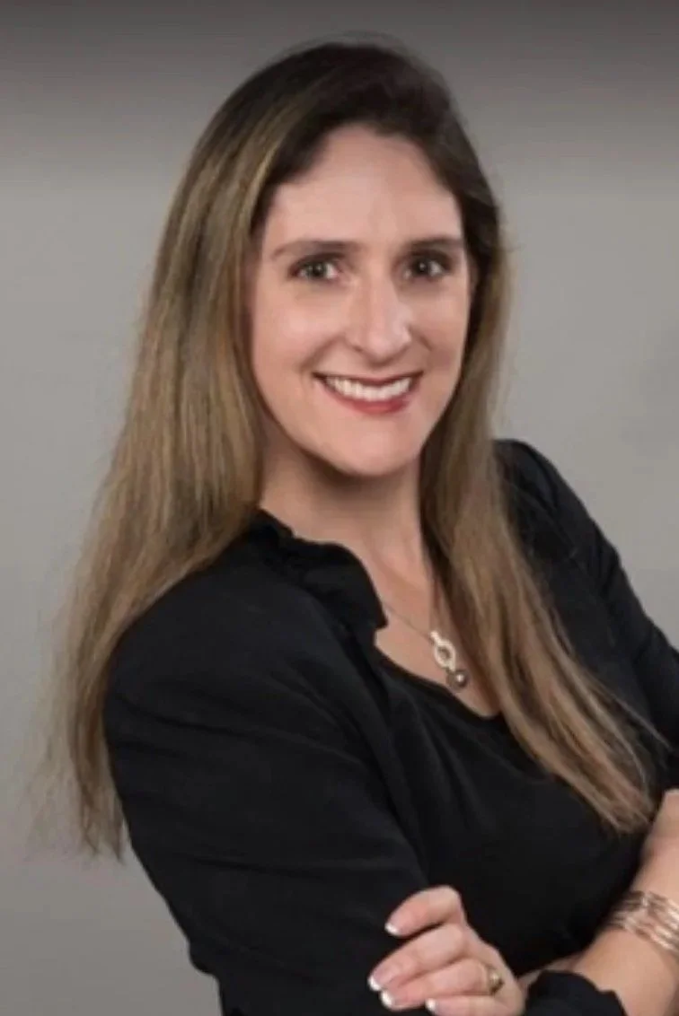 A woman with shoulder-length light brown hair smiling, wearing a black top and a necklace, with arms crossed, in front of a plain gray background.