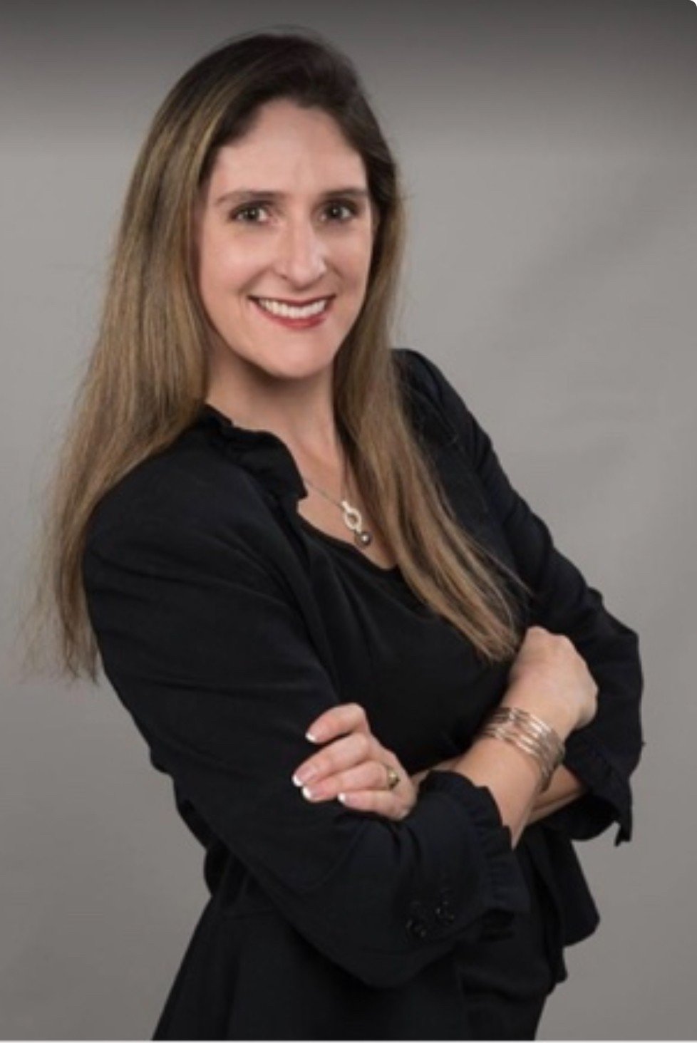 A woman with long, light brown hair smiling, wearing a black blouse, with arms crossed, against a gray background.