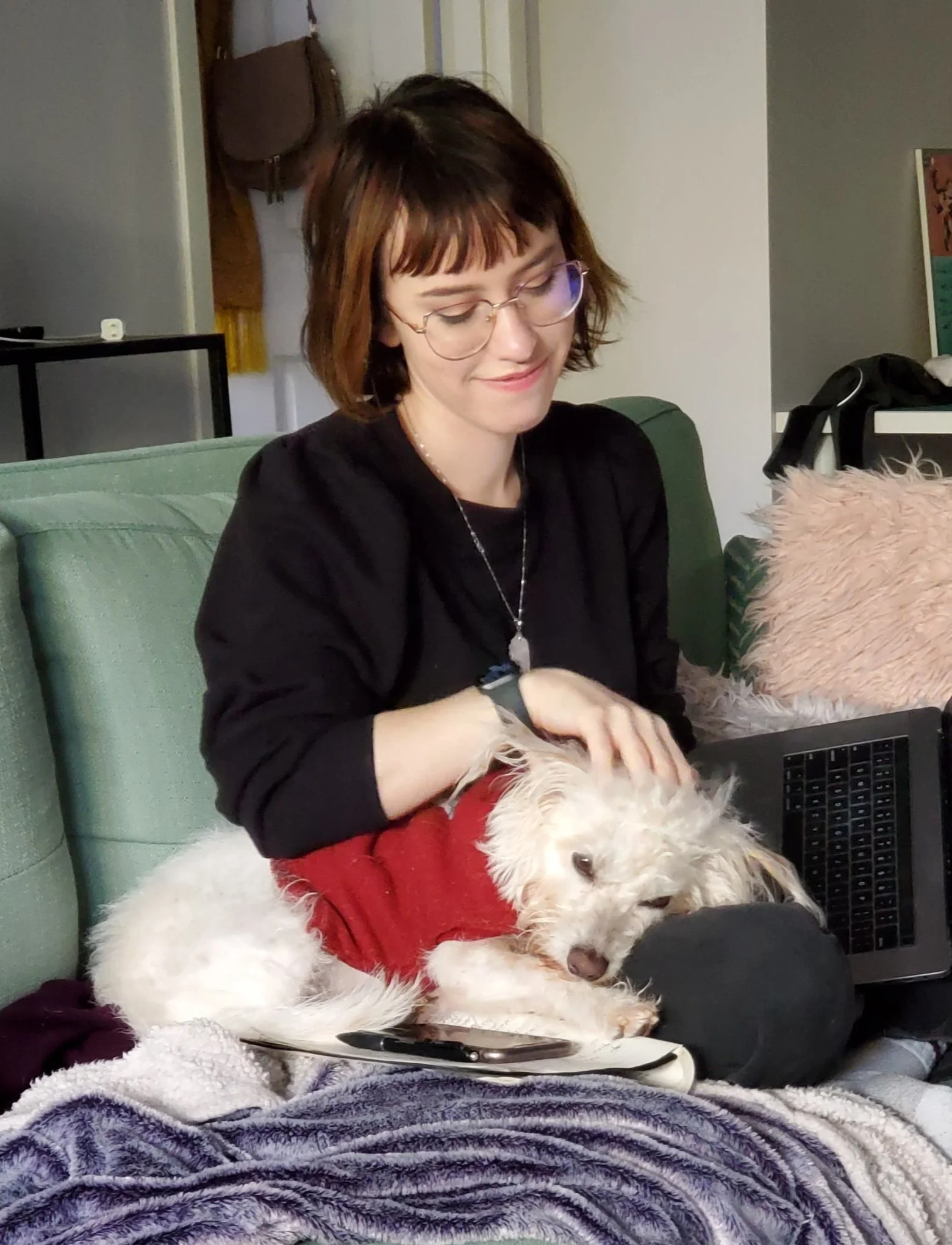 A person in glasses petting a white dog wearing a red sweater, sitting on a green couch with a laptop nearby.