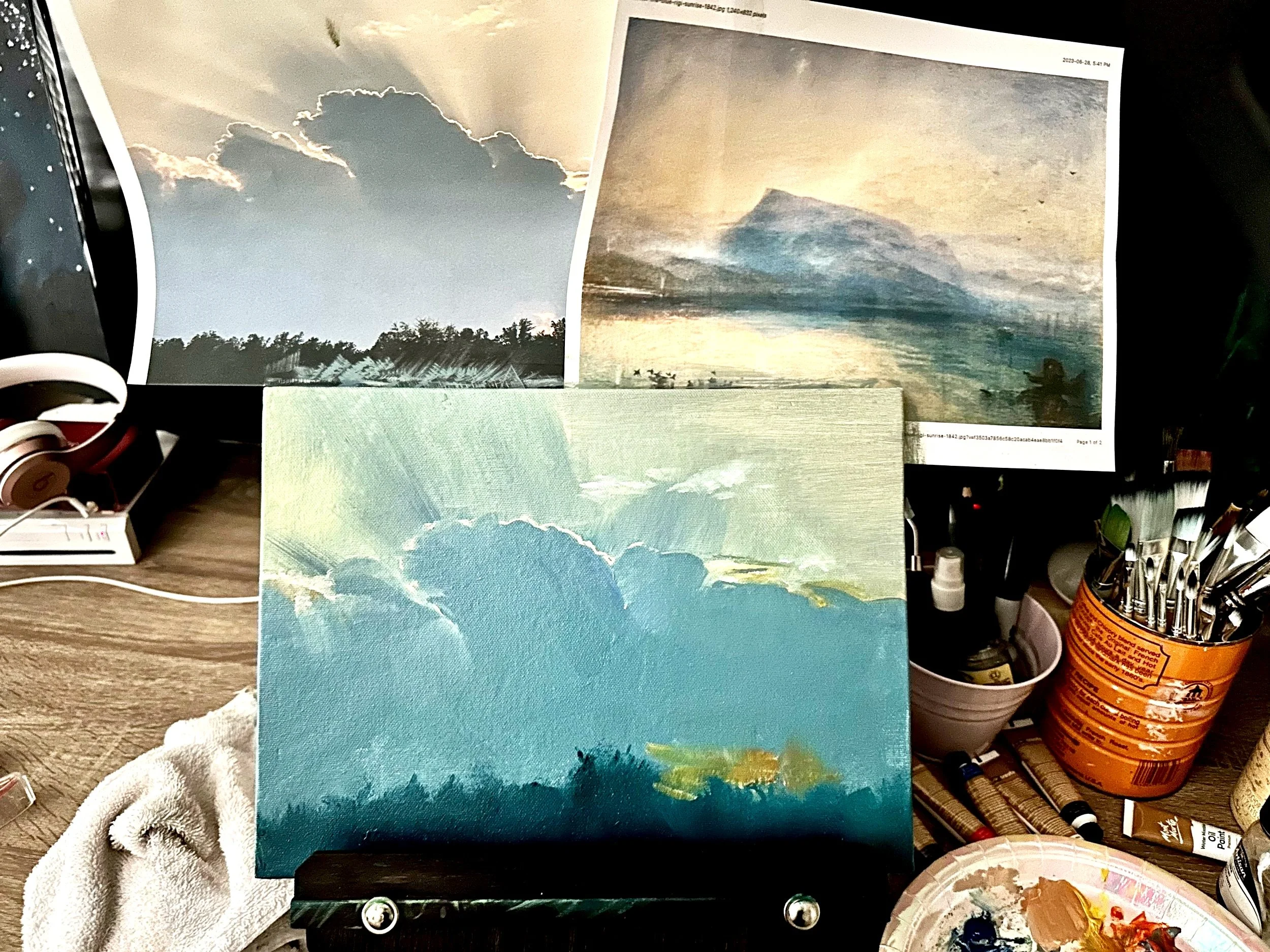 Art studio setup with a blue and green landscape painting on an easel, a palette with mixed paints, paintbrushes in a can, and inspirational images on a cluttered desk.