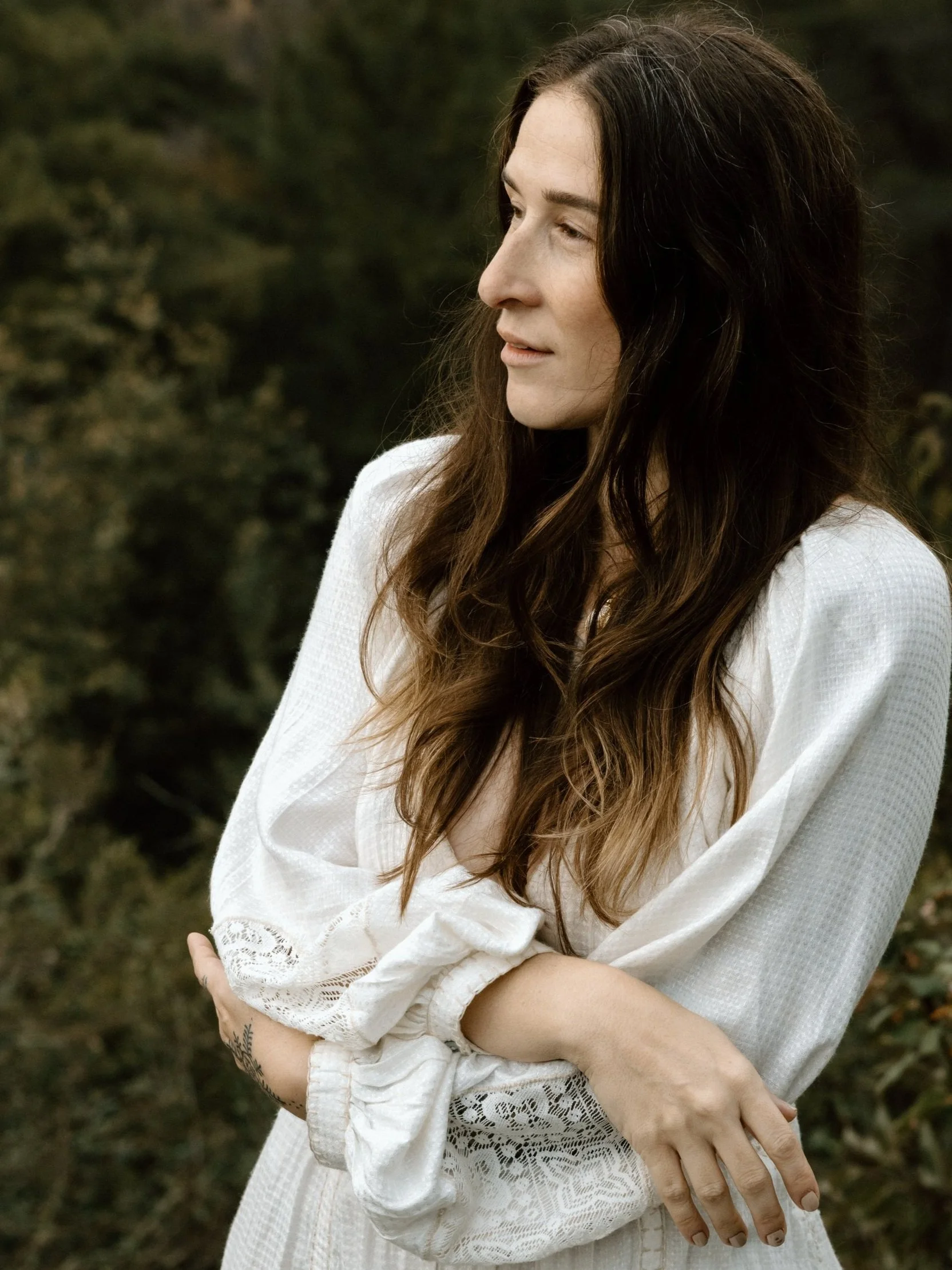 A woman with long, wavy brown hair wearing a white long-sleeve dress with lace details, standing outdoors with her arms crossed, looking to the side, in front of blurred greenery.