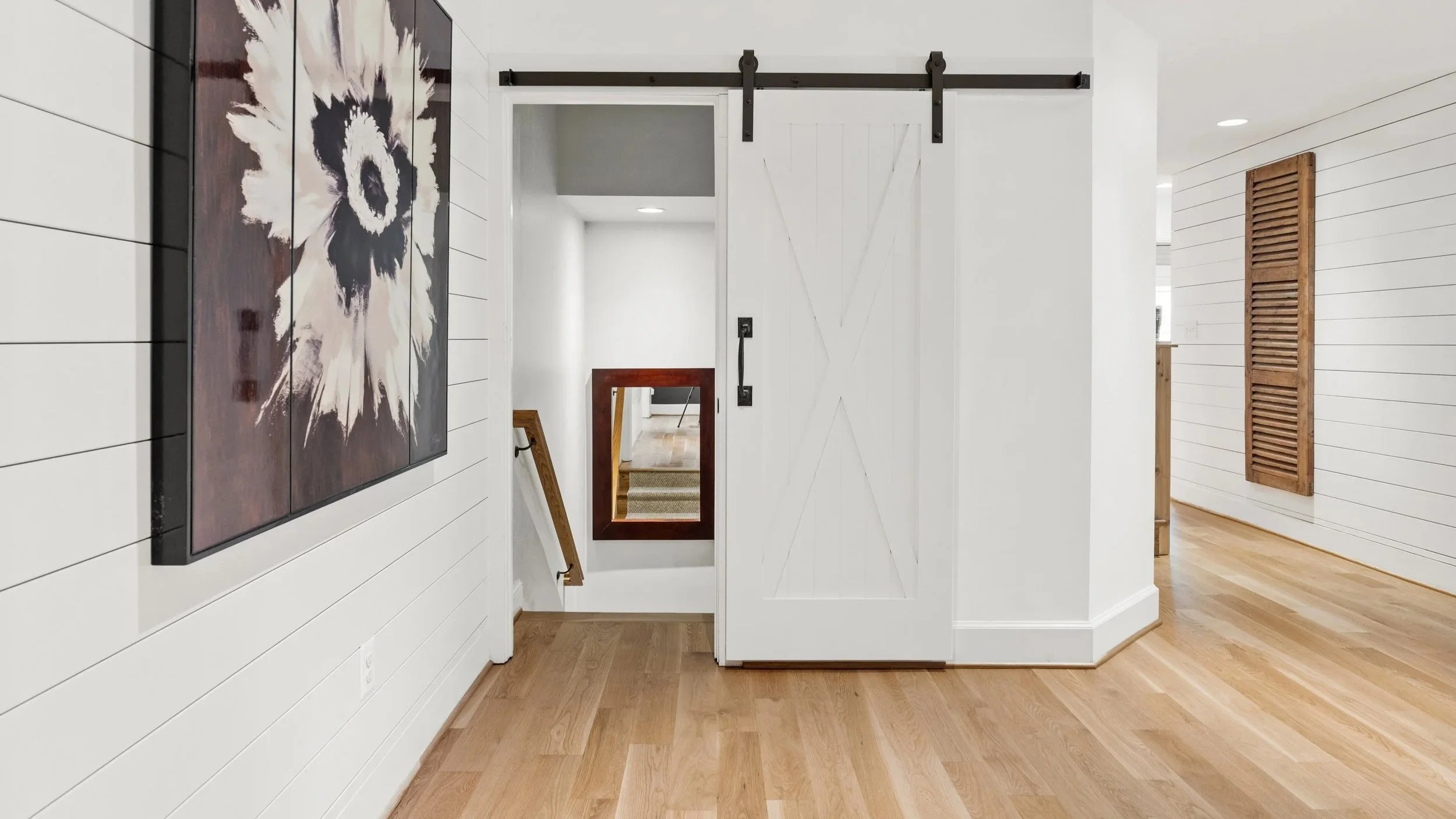 Interior of a modern home hallway with light hardwood floors, white shiplap walls, a large floral artwork, a sliding barn door, and a staircase with a mirror.