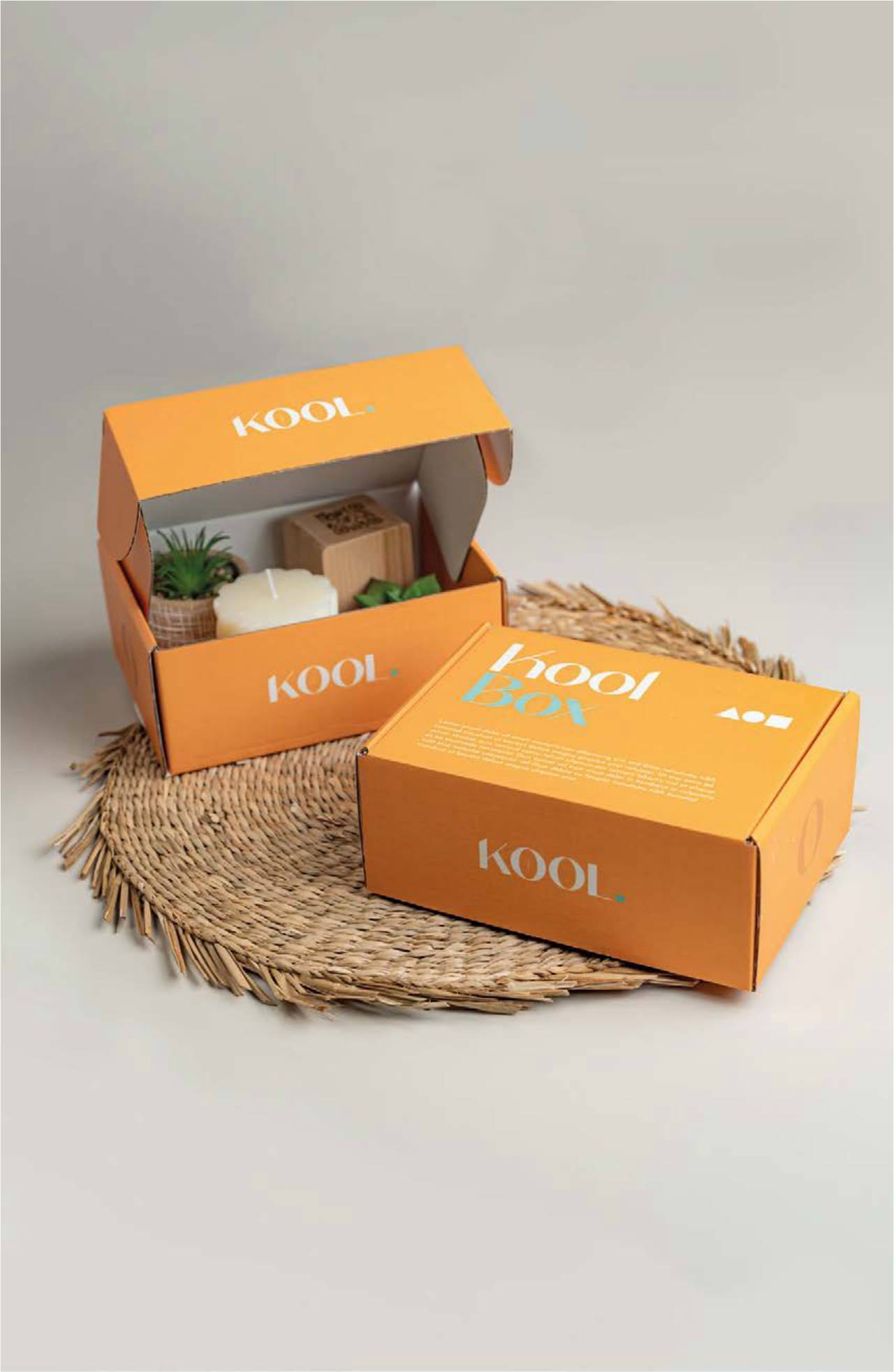 Une boîte orange de la marque KOOL ouverte, contenant différents objets décoratifs comme une bougie, une plante en pot, et un petit cube en bois, posée sur une nappe ronde en corde sur fond blanc.