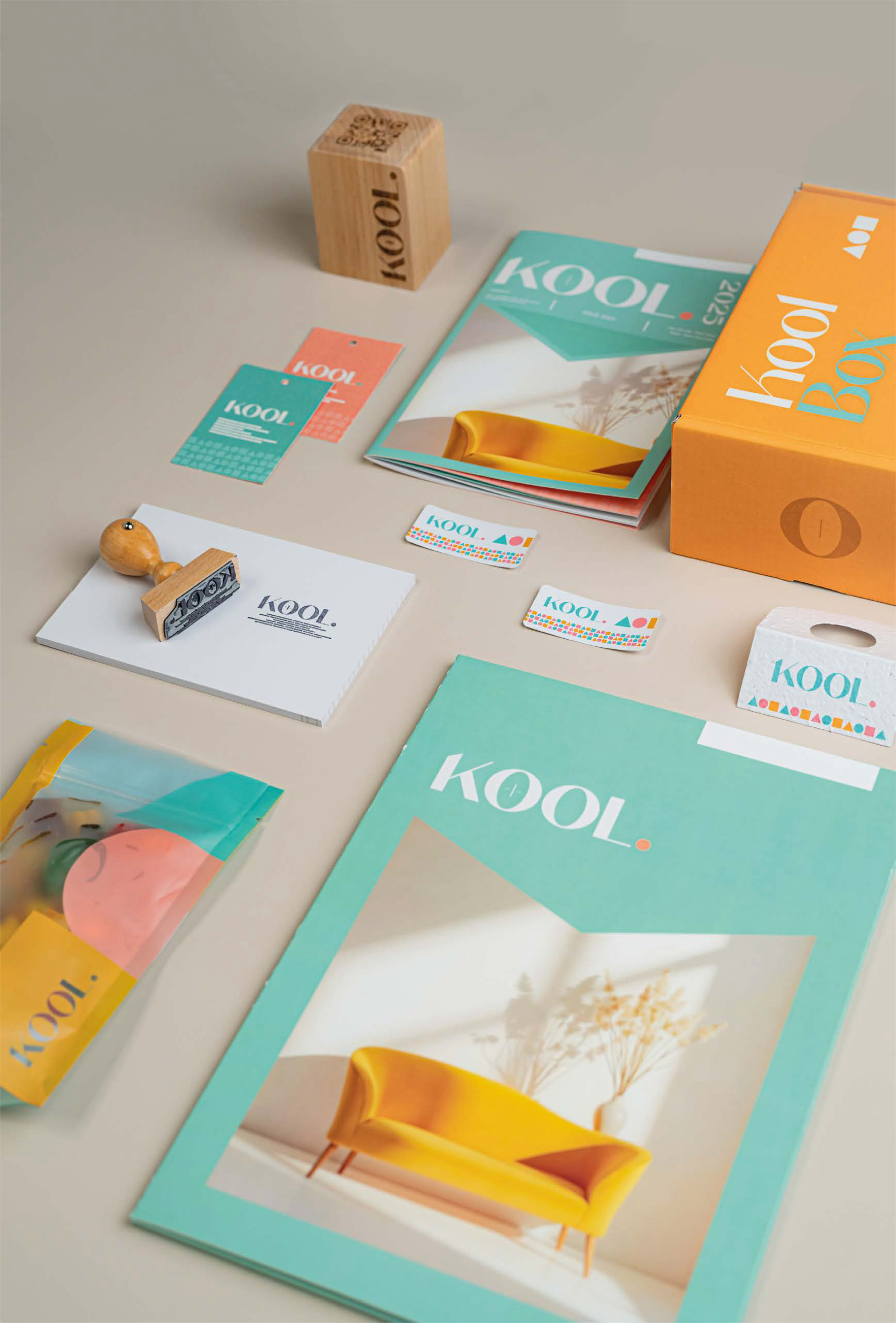 Une mise en page de matériel promotionnel pour Kool, avec une boîte orange, des magazines bleus et roses, des étiquettes colorées, et des éléments décoratifs comme un cube en bois et une chaise jaune.