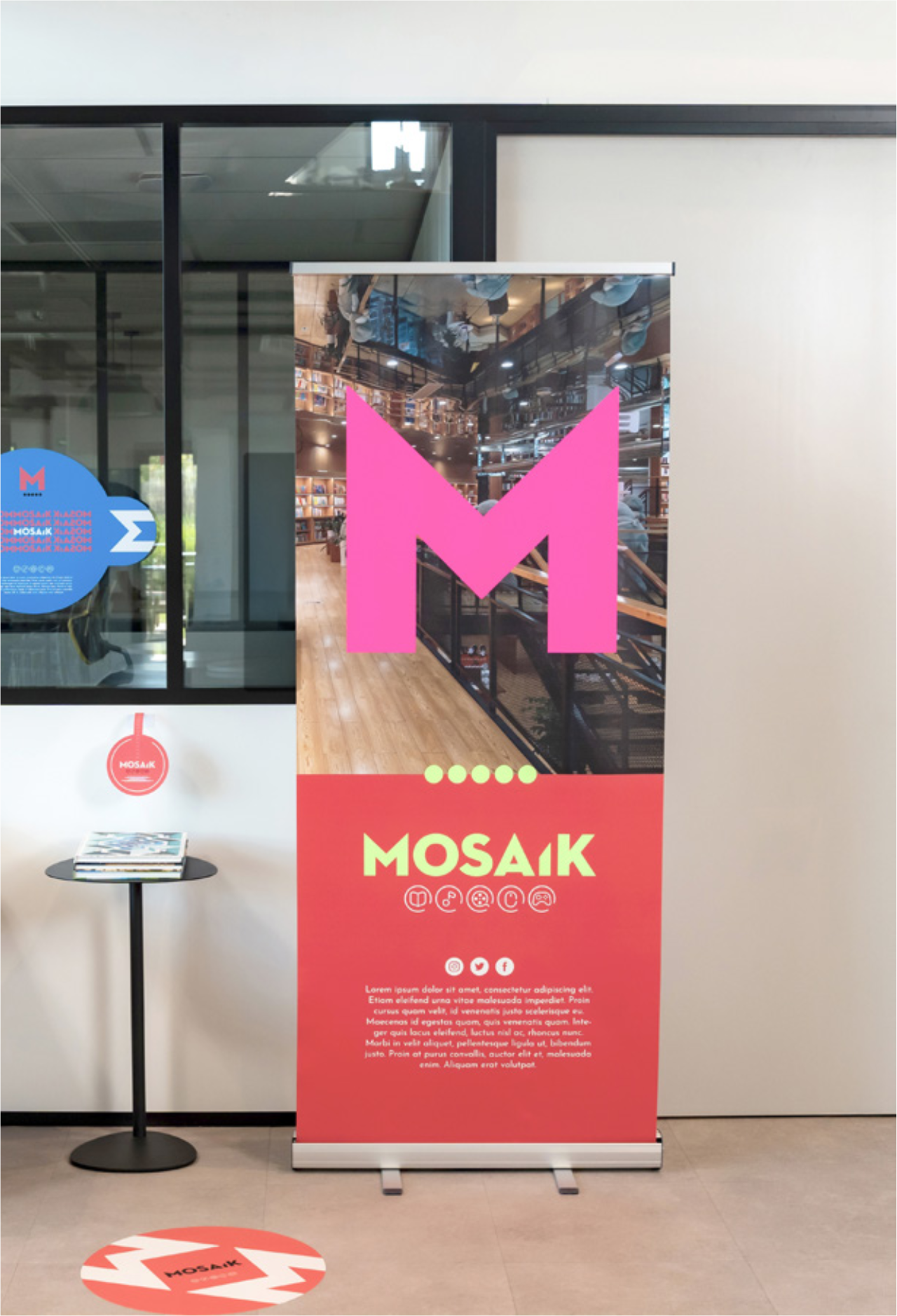 Un panneau d'affichage rouge et rose avec le logo Mosaik, situé à l'intérieur d'un bâtiment, à côté d'une petite table ronde avec des magazines dessus, et un cercle au sol avec le même logo.