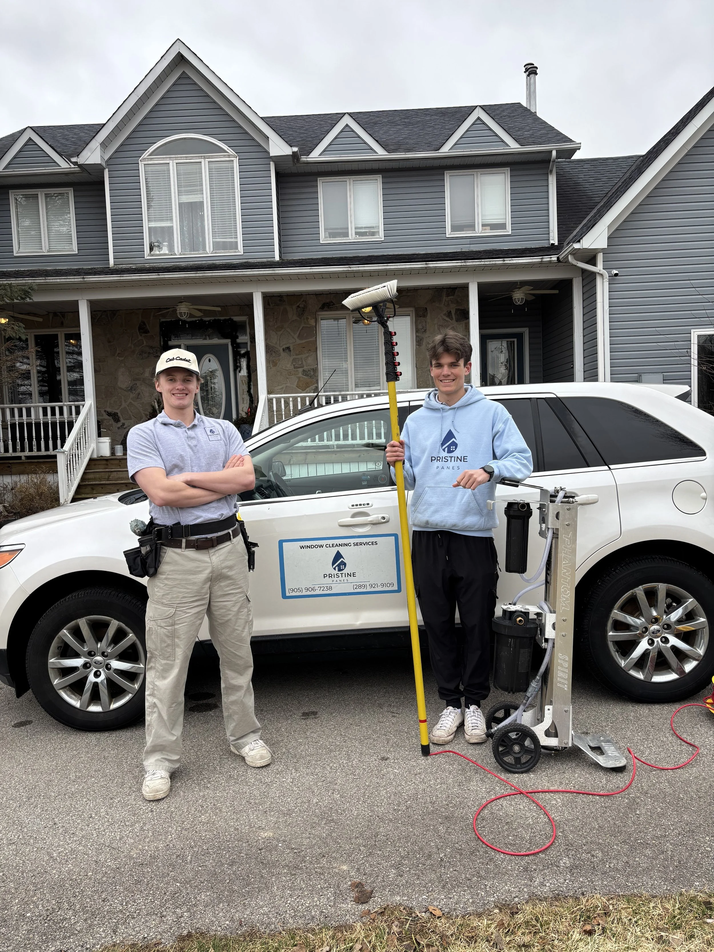 Two technicians from Pristine Window Cleaning Services standing in front of a house with a white car, holding cleaning equipment.