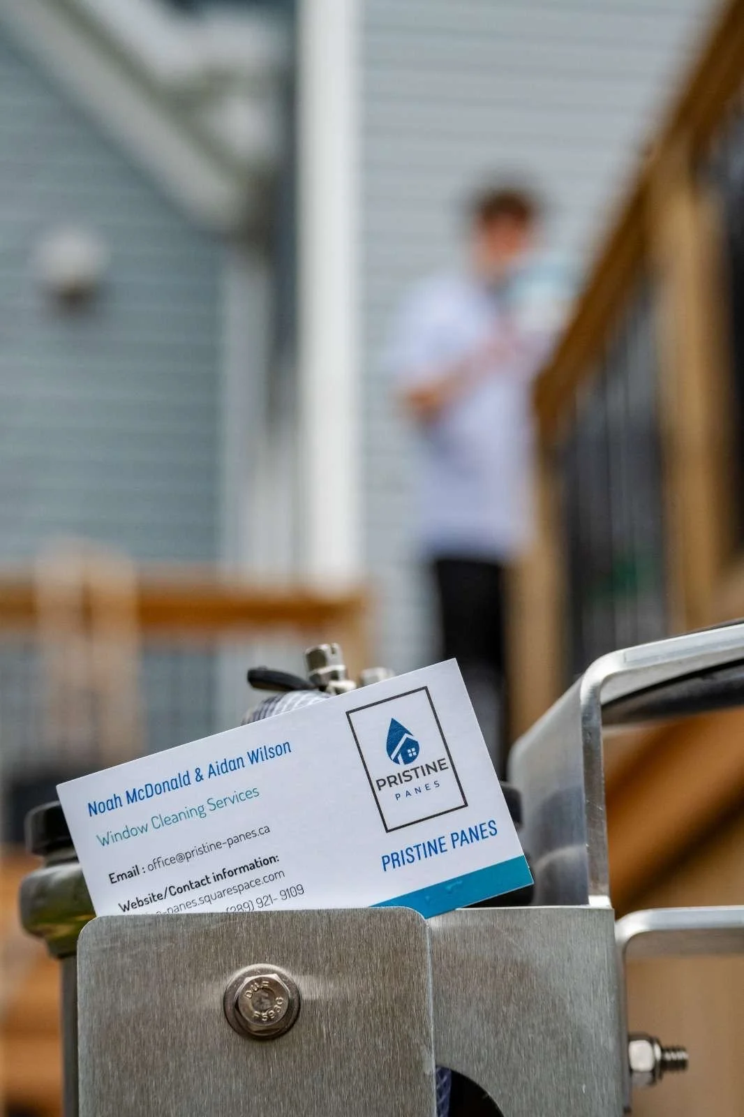 Close-up of a business card for Noah McDonald & Aidan Wilson, Window Cleaning Services, placed in a metal holder attached to cleaning equipment with a blurred person in the background.