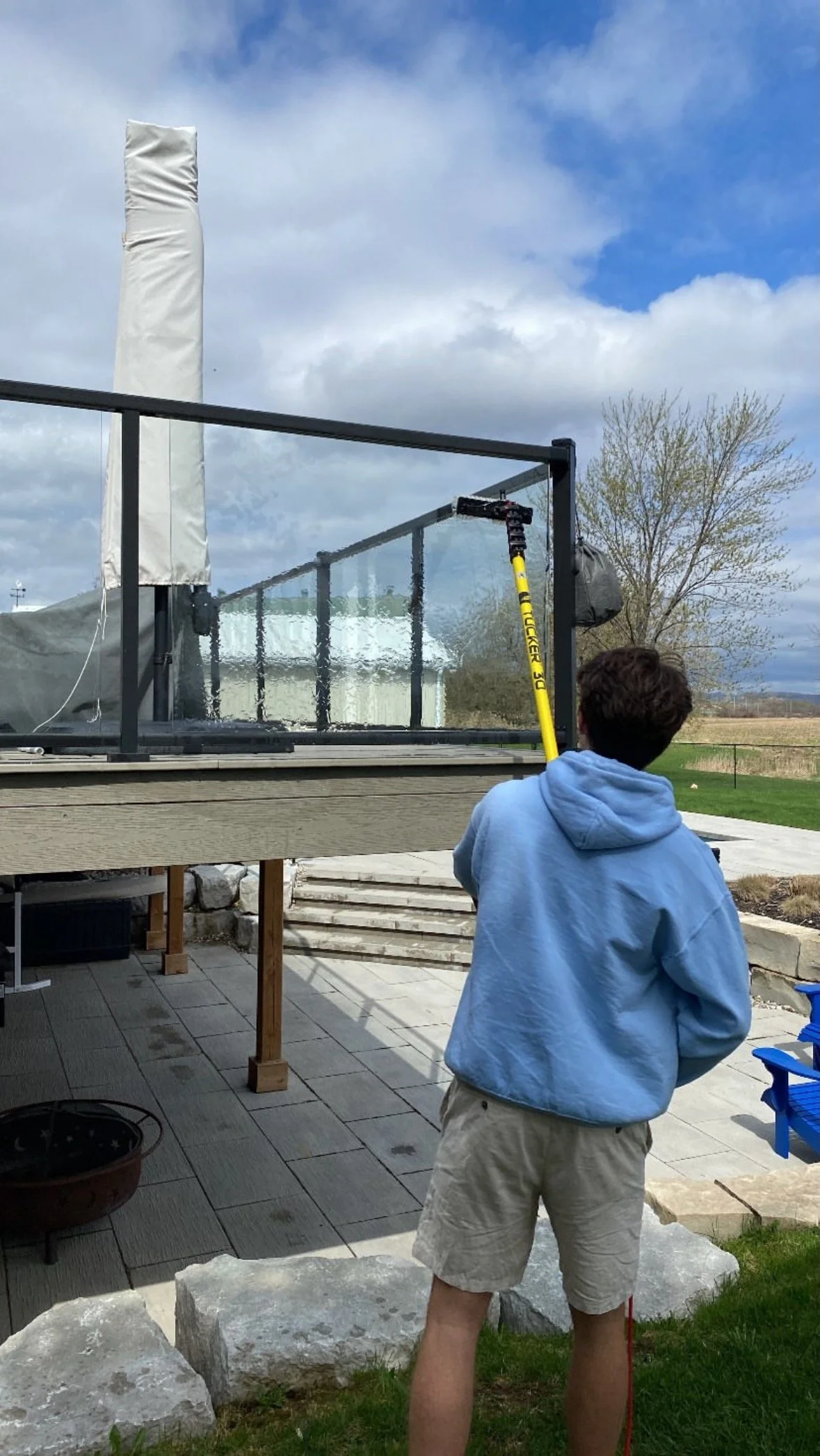 A person in a light blue hoodie and beige shorts cleaning a large glass window on a patio with a long yellow tool, with cloudy sky and trees in the background.
