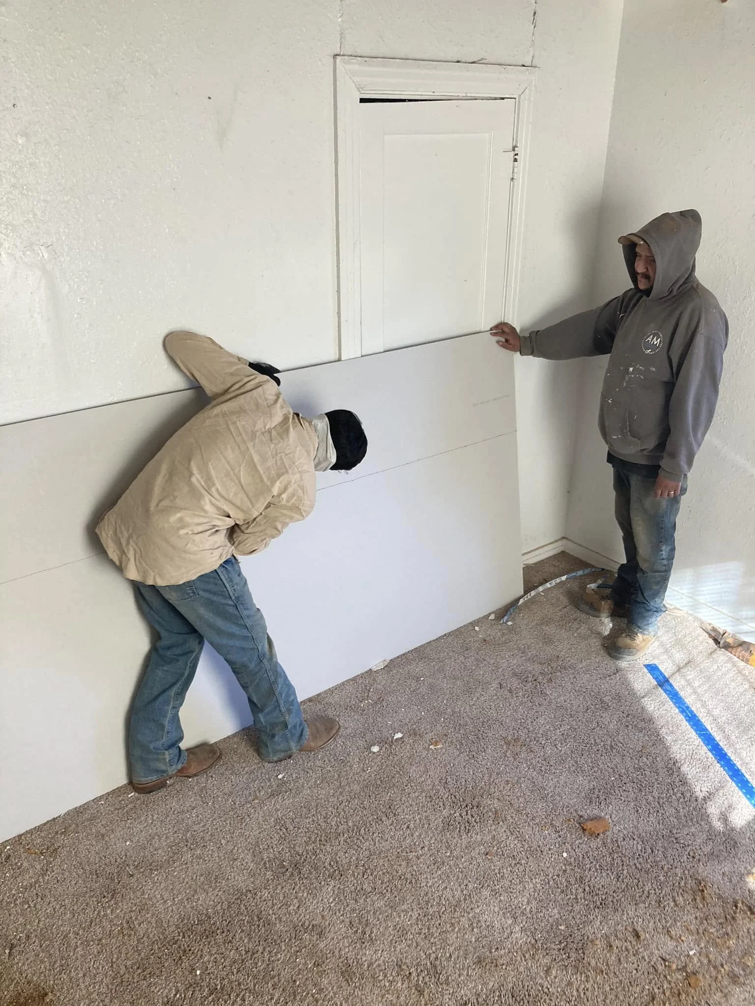 Two workers installing drywall on a wall in a room with a beige carpet, one holding a drywall panel and the other guiding it into place.