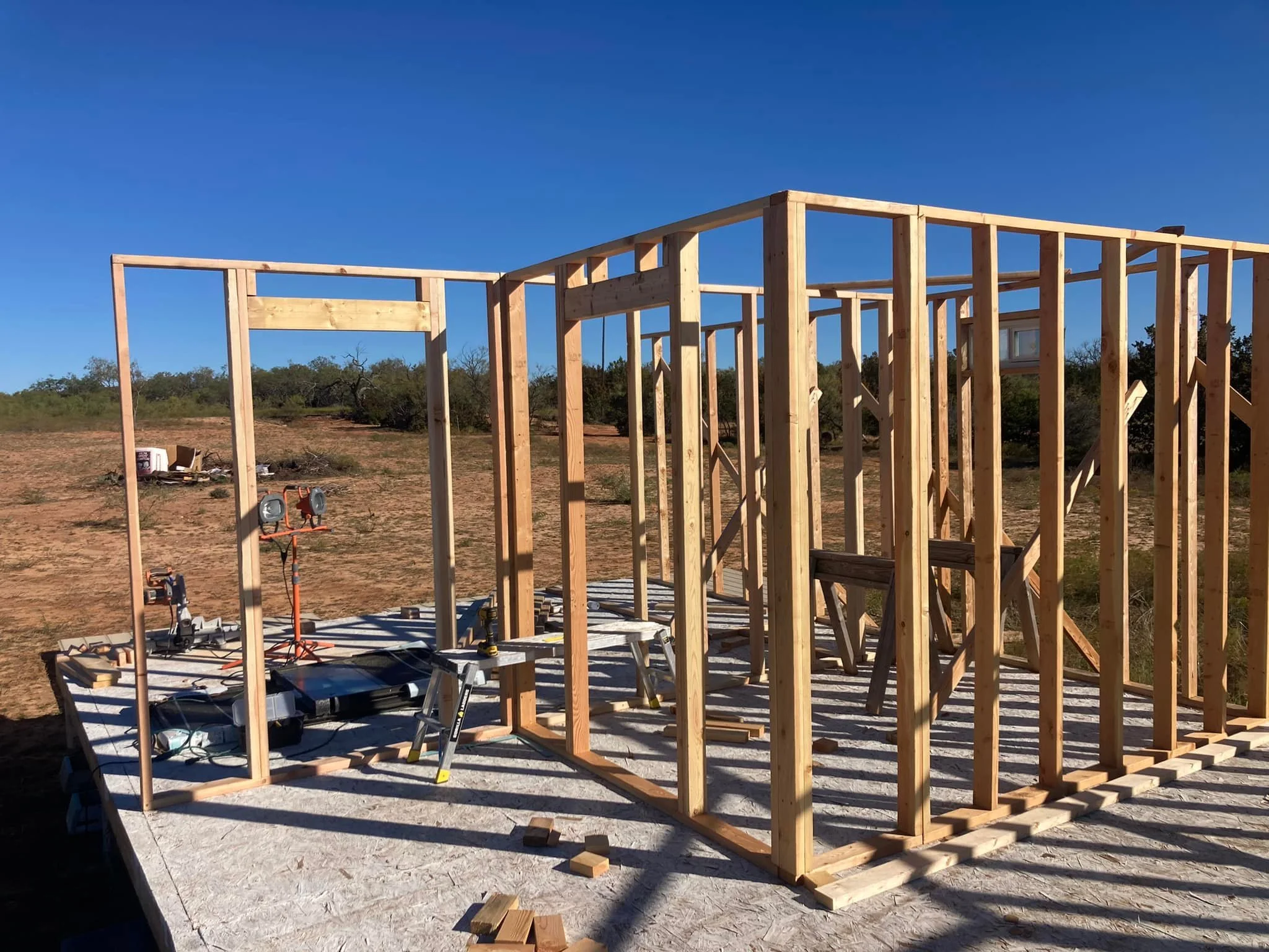 Wooden framing of a new building under construction on a concrete slab with a clear blue sky and some distant trees.