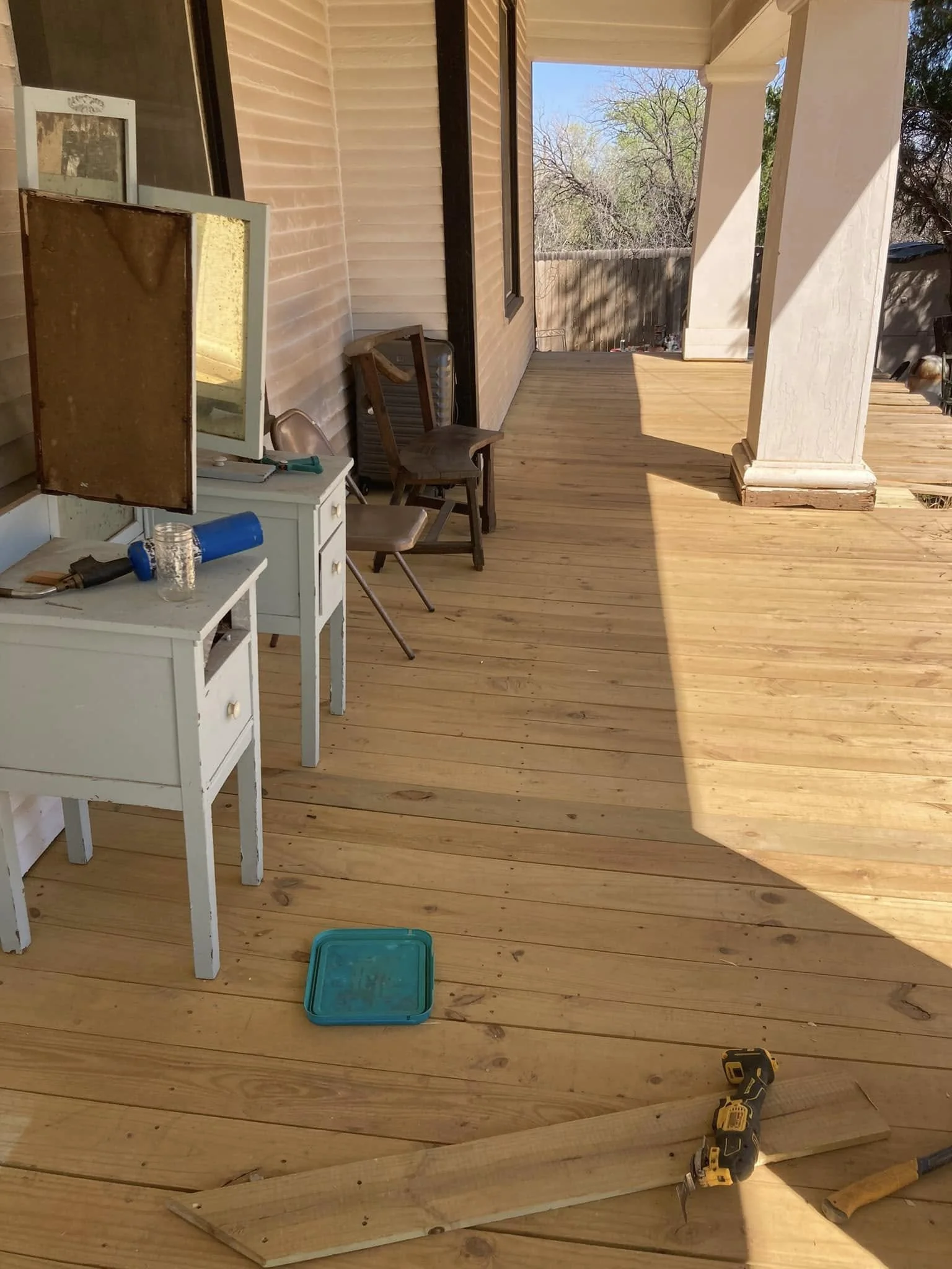 This image shows a partially built wooden deck with construction tools and furniture. There are two small nightstands or dressers, a mirror, chairs, a blue container lid on the floor, and some construction materials including a yellow cordless drill 