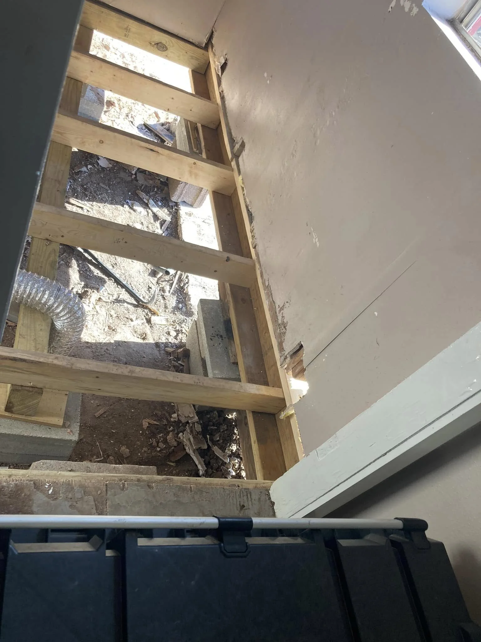 Interior view of a construction site showing the framing for a new wall, with exposed wooden studs, insulation vents, and a dirt floor.