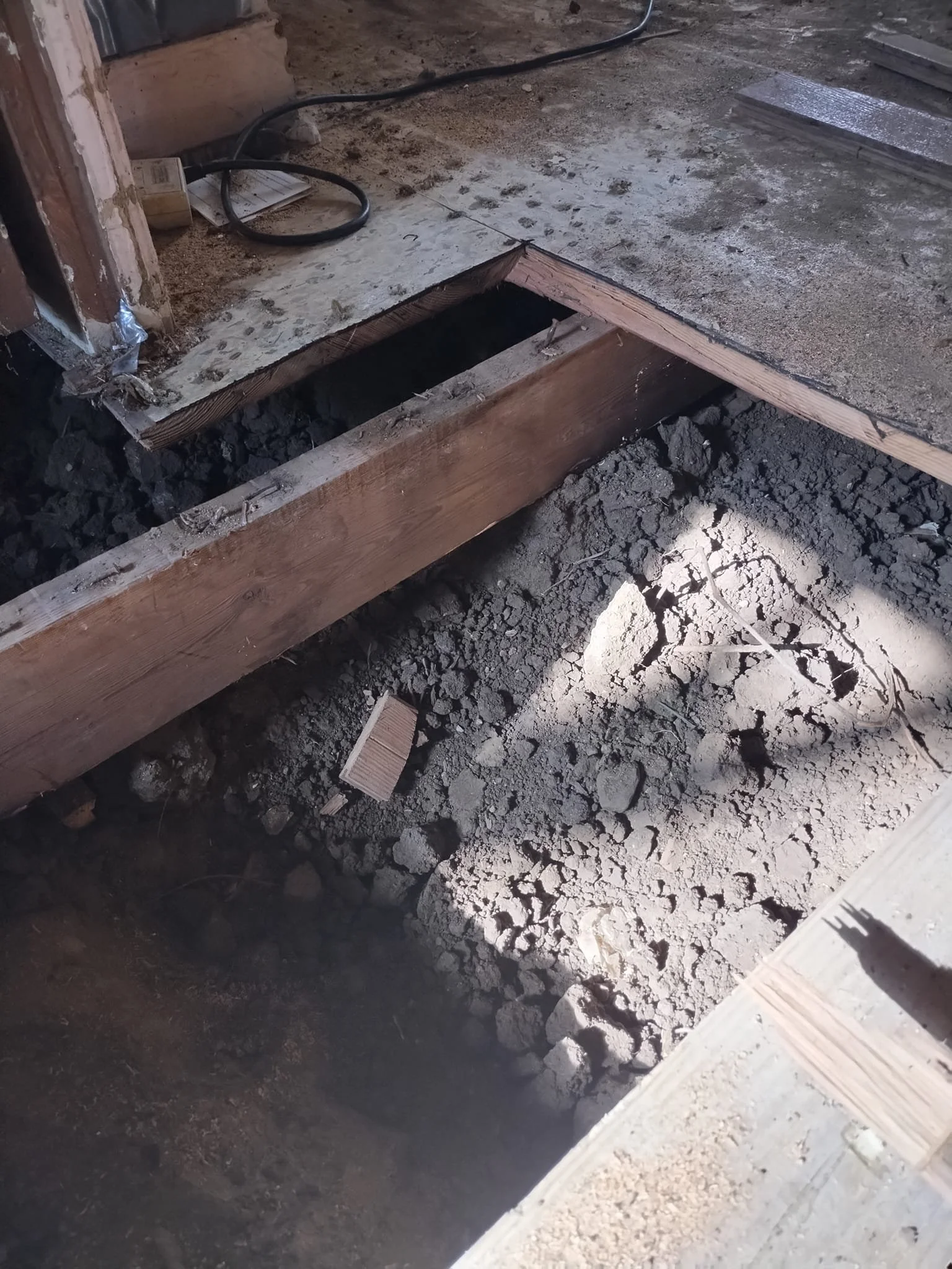 View of a construction site with dirt and debris, showing a section of floor under renovation with a large hole and exposed wooden framing.