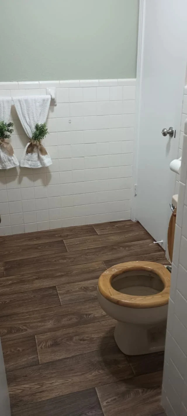 A bathroom with a toilet that has a wooden seat, white tiled walls, a white door, and wood-look tile flooring, with two small towels hanging on a rail with plant accents.