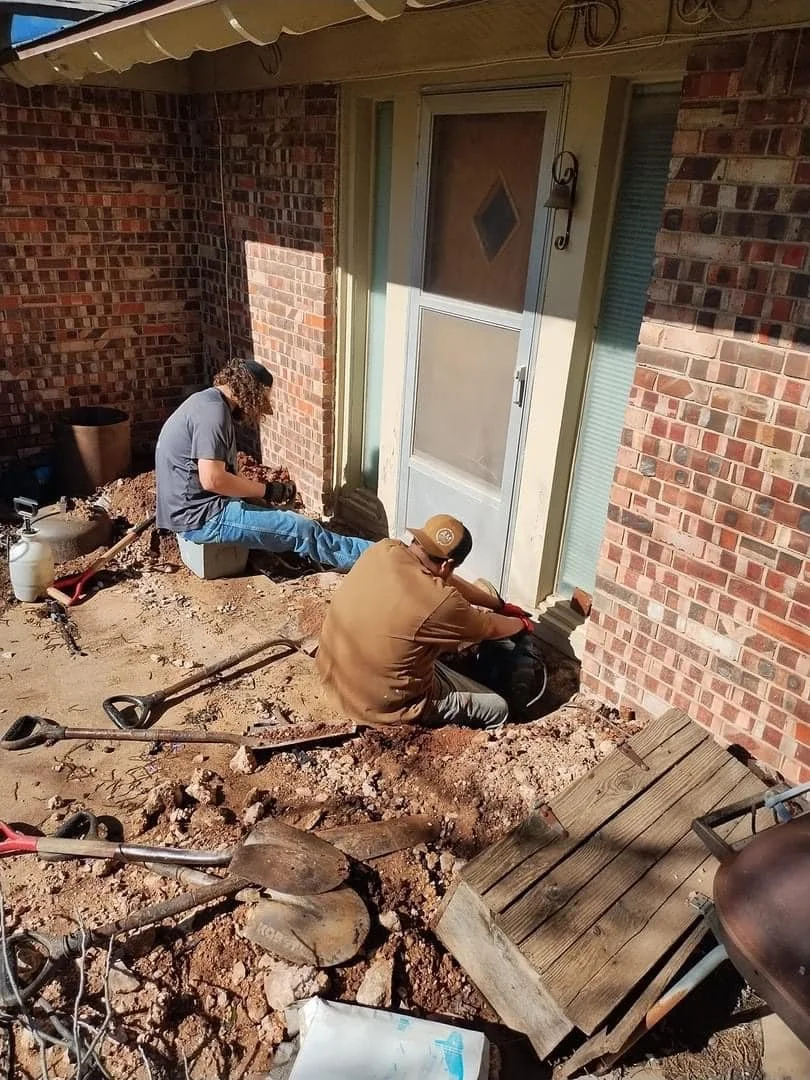 Two people working on a construction or repair project outside a house, digging in the dirt near the door, with gardening tools and a wooden ramp on the ground.