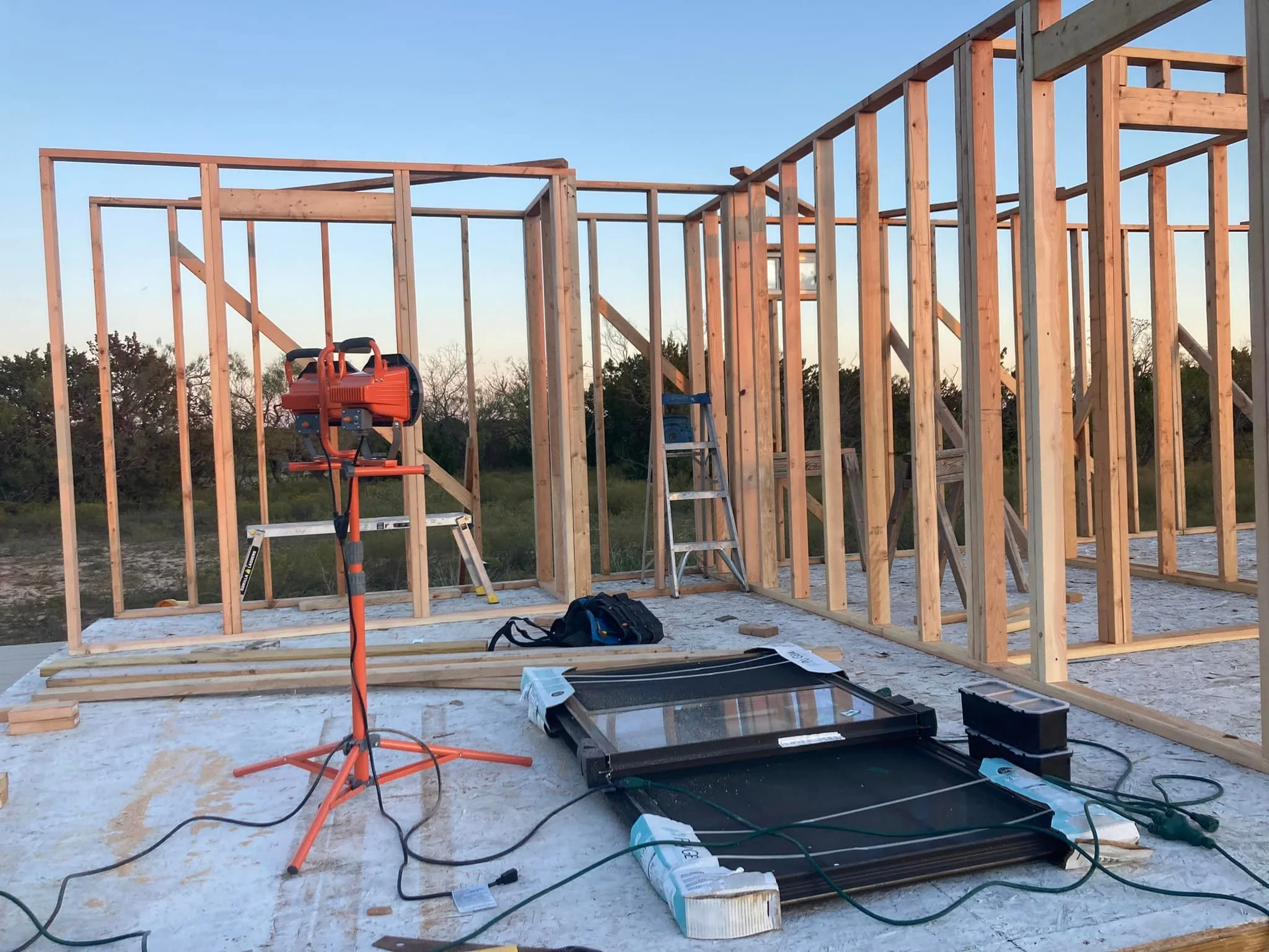 Wooden framing of a house under construction with construction tools and materials on the ground, including a ladder, power saw, and solar panel, outdoors during daylight.