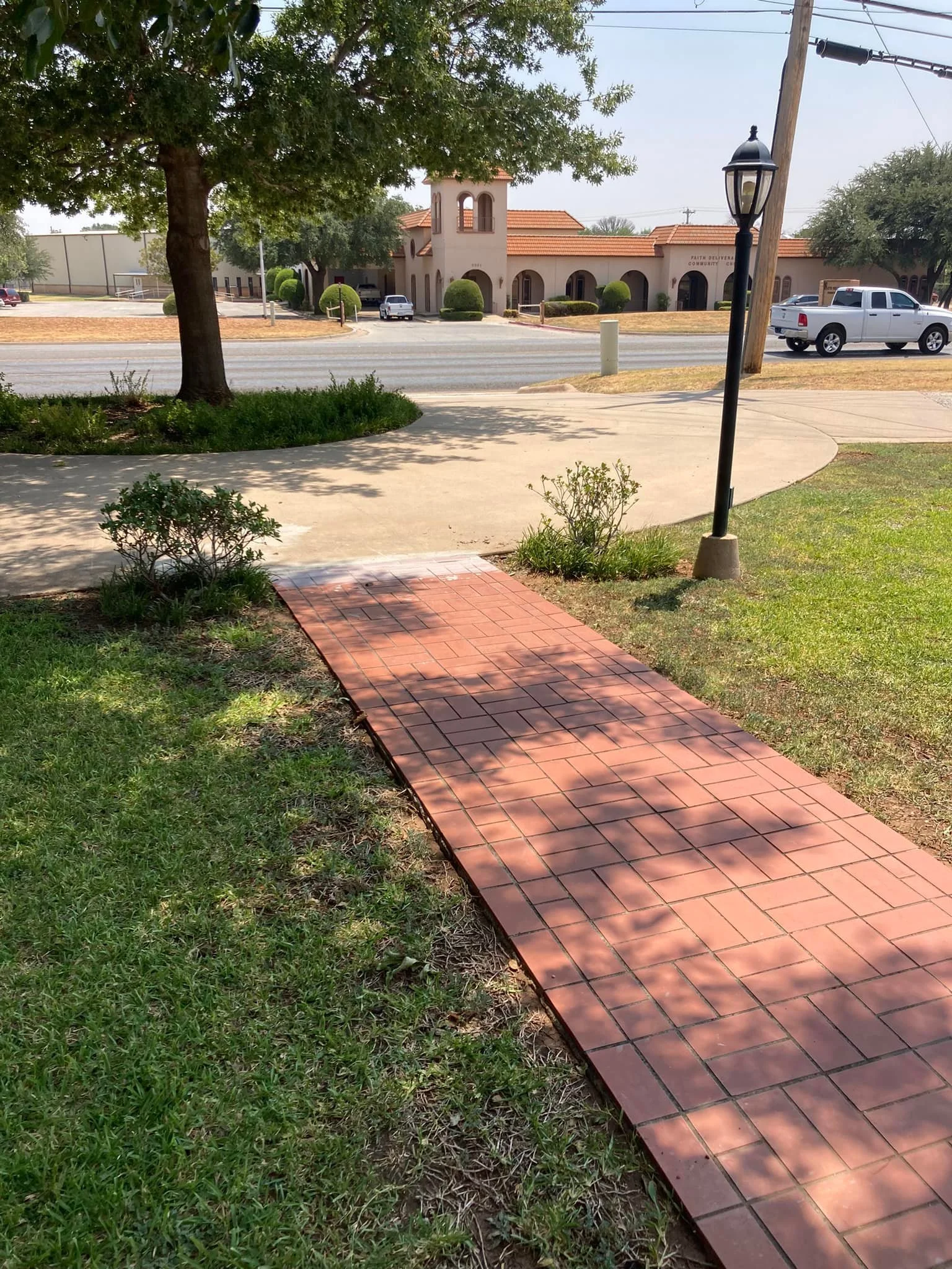 A sidewalk with red bricks curves from the foreground to the middle ground, with grass and bushes on either side. There is a street in the background with vehicles and a building with a tower and red tile roof. A street lamp and a lamppost are also v