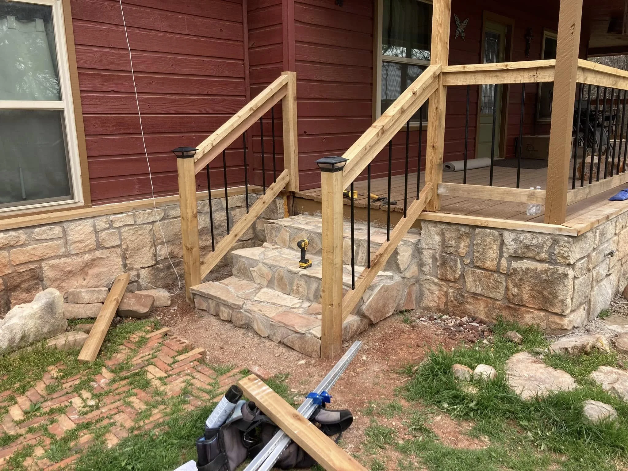 The image shows a partially constructed outdoor wooden staircase with black metal balusters and stone steps, attached to a red wooden house with a stone foundation. There are construction tools on the ground, including a drill, a saw, and some bottle
