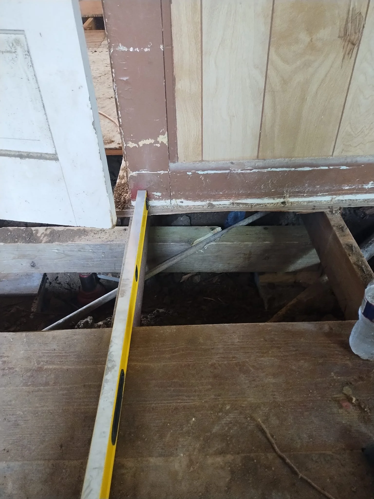 Close-up of a construction site showing a level tool resting on a wooden plank, with a partially installed door frame and wooden wall panels in the background.