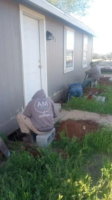 Two workers installing or repairing a sidewalk beside a house with gray siding and white-framed windows