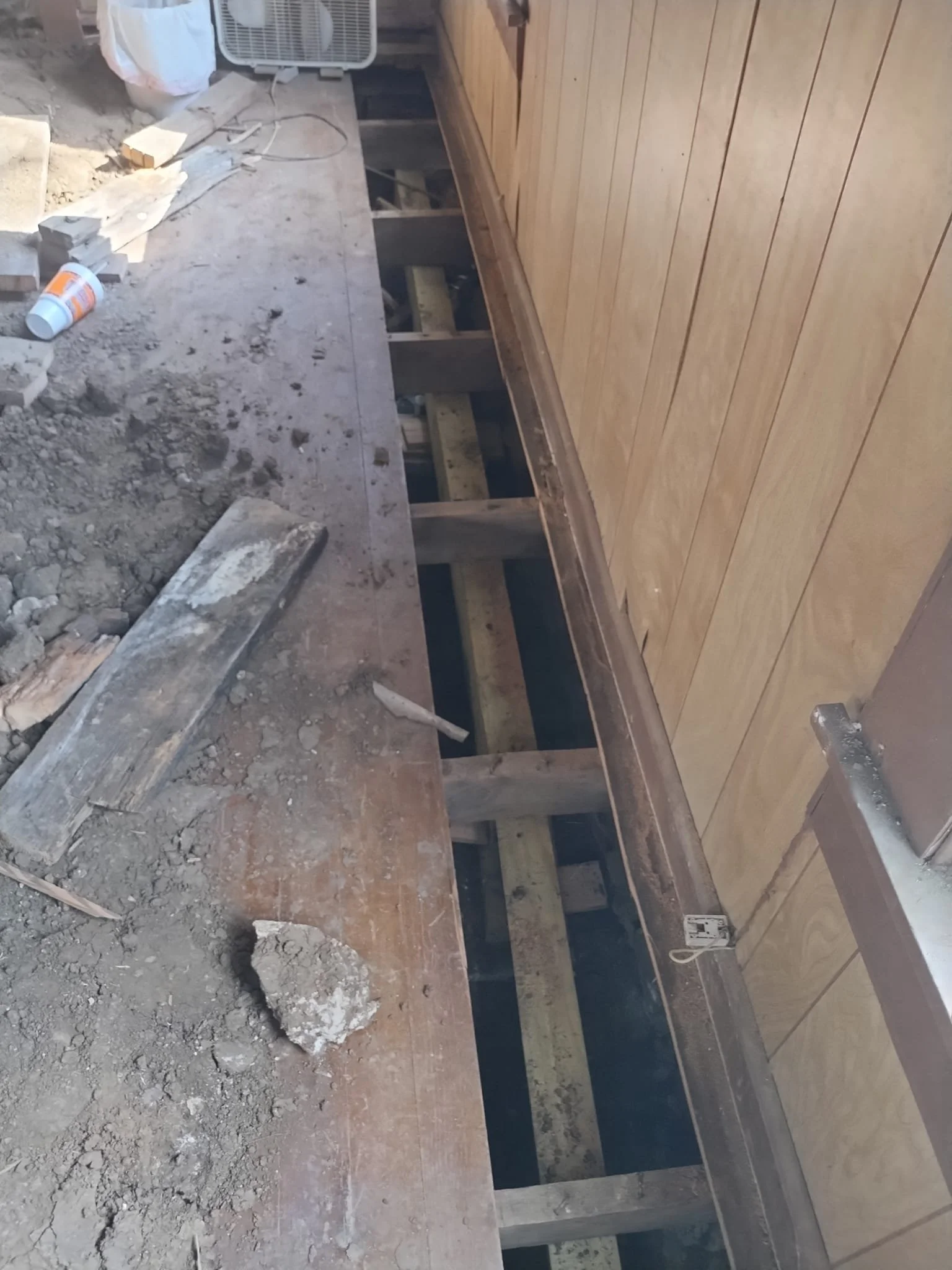 Construction scene showing a wooden floor or platform being built with visible support beams; tools, debris, and supplies scattered around.