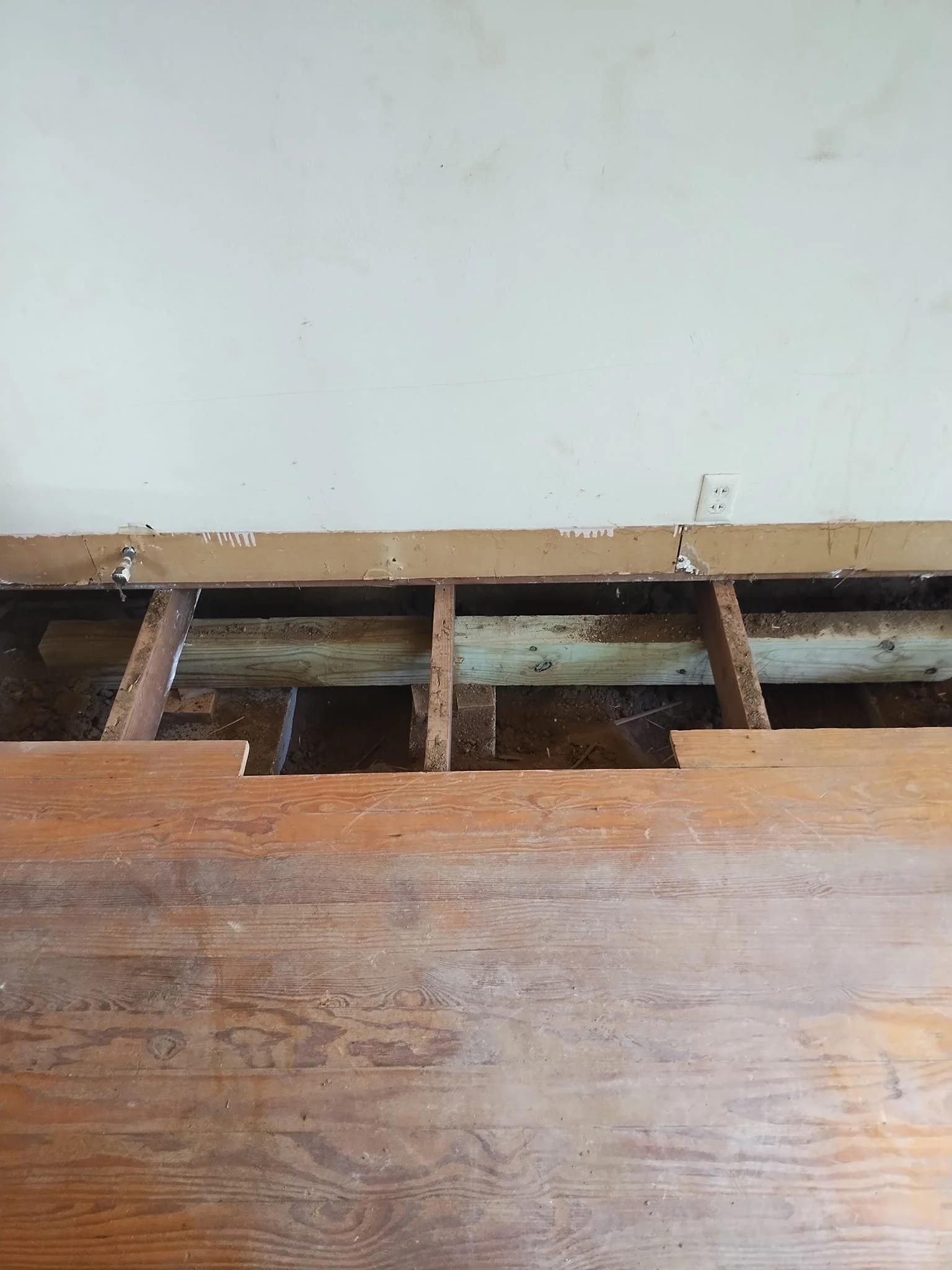 A room with the flooring partially removed, exposing wooden floor joists and dirt underneath. A wall with an electrical outlet is visible in the background.
