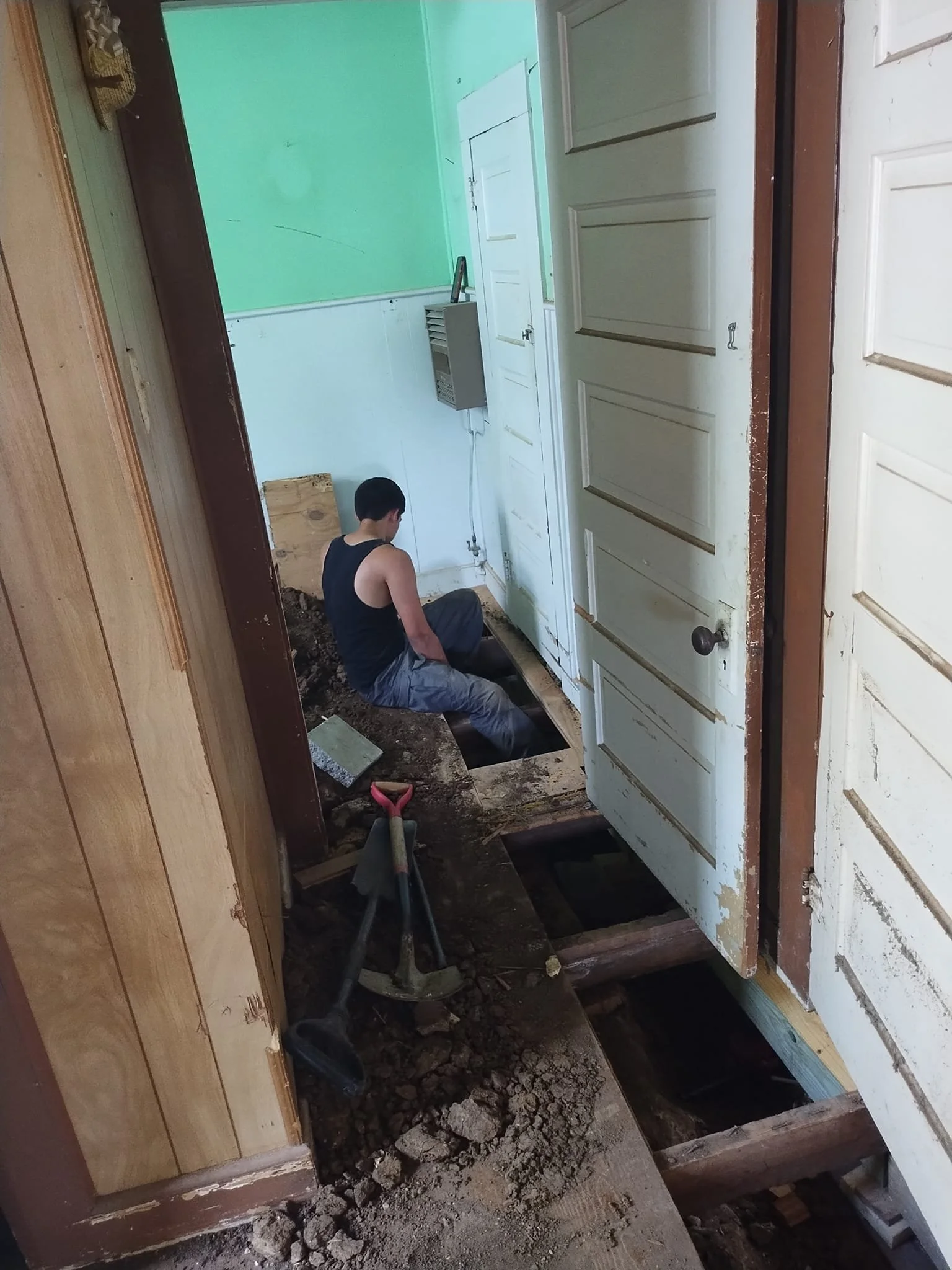 A person working on a home renovation project, sitting on the floor of a room with exposed floor joists, surrounded by tools and dirt, with a partially removed floor.