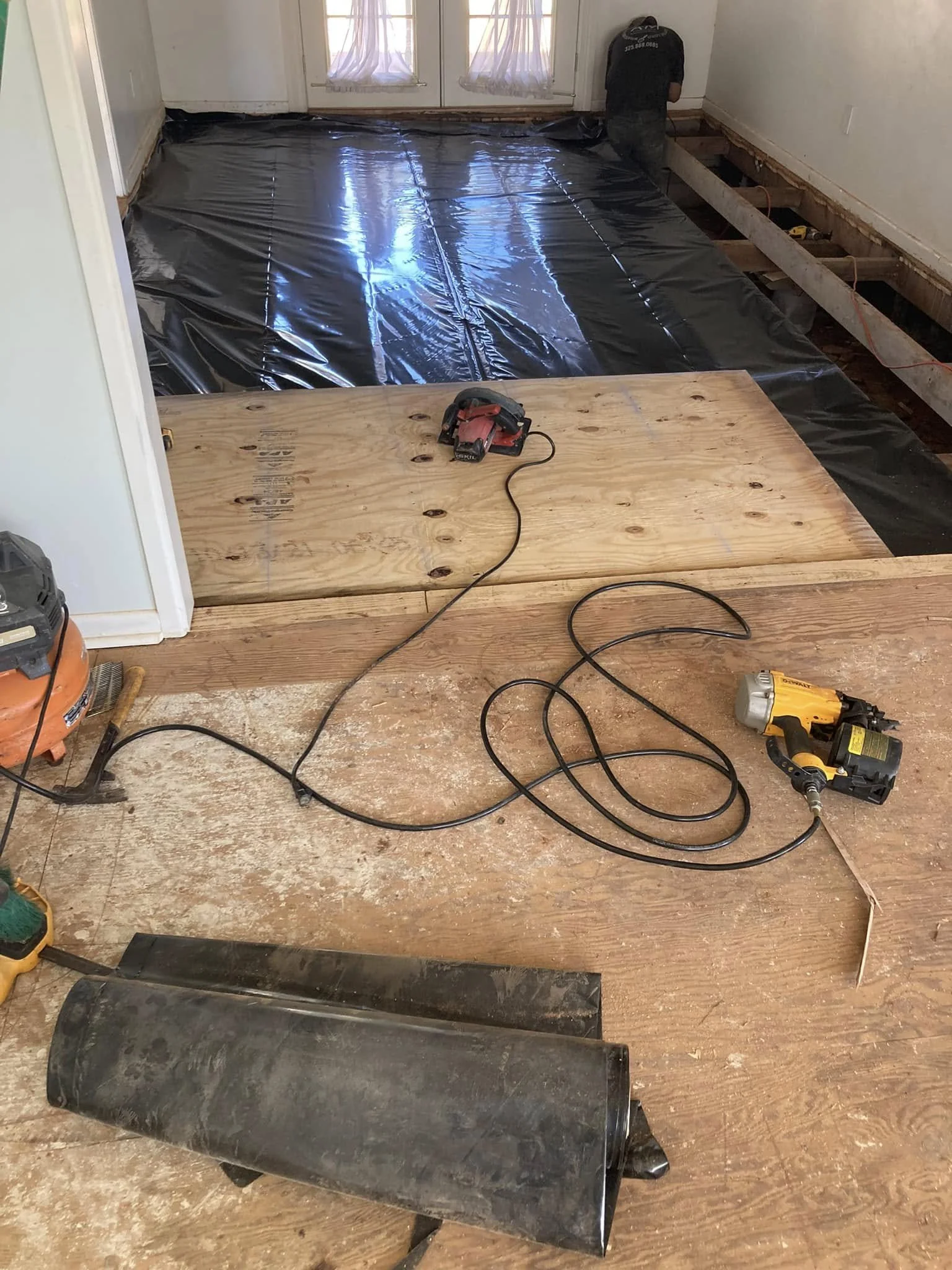 A room under renovation with a black plastic sheet on the floor, construction tools, and a person working near a door with curtains. New plywood flooring is being installed.