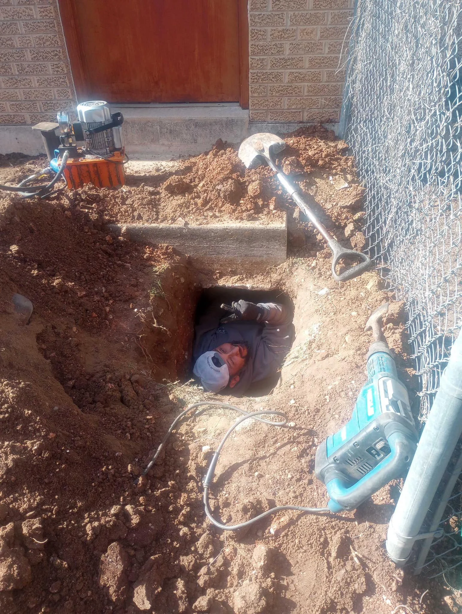 Construction worker lying in a large rectangular hole in the ground, with a shovel nearby, surrounded by dirt and construction tools.