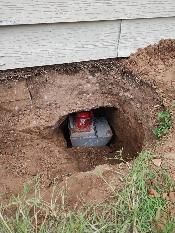A buried utility box with a red cap in a hole near the foundation of a building with white siding.