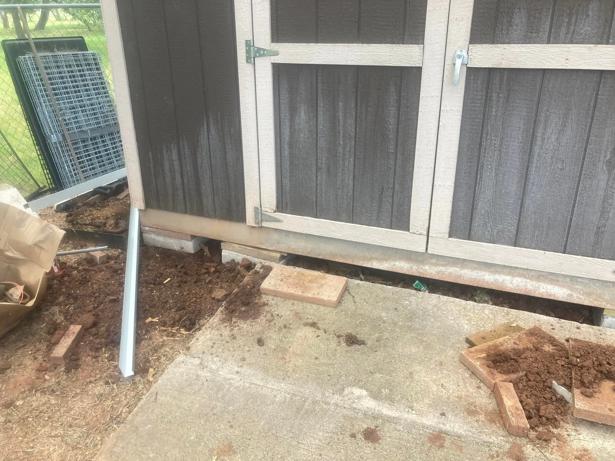 Construction or repair work in progress on a shed or small building, with exposed dirt and bricks at the base and tools nearby.