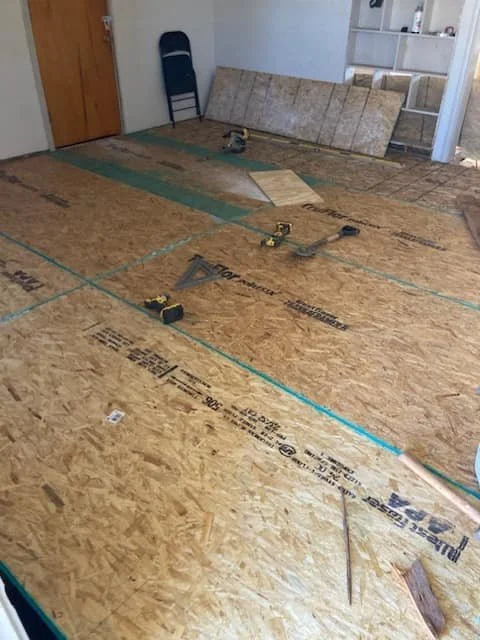 Room under construction with plywood subflooring, power tools, and blue measuring lines, indicating flooring installation in progress.