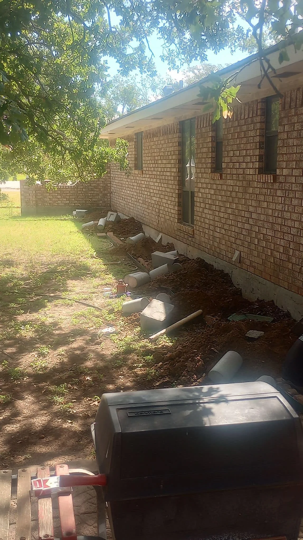 Backyard with ongoing exterior wall construction, with PVC pipes along the ground, dirt, and a grill in the foreground.