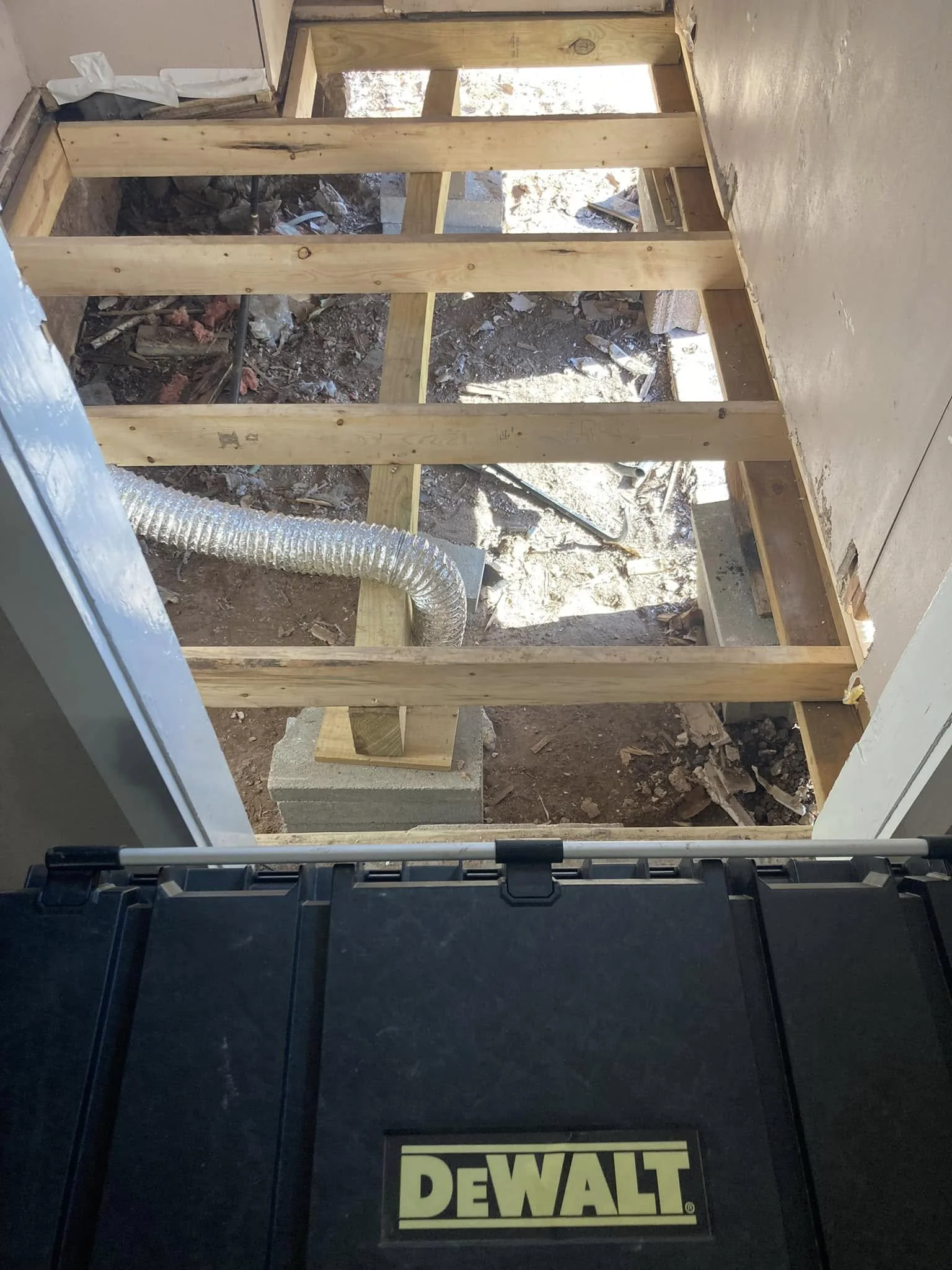 View from doorway showing a construction site with wooden framing for a floor, exposed dirt, a flexible duct pipe, and construction tools and debris.