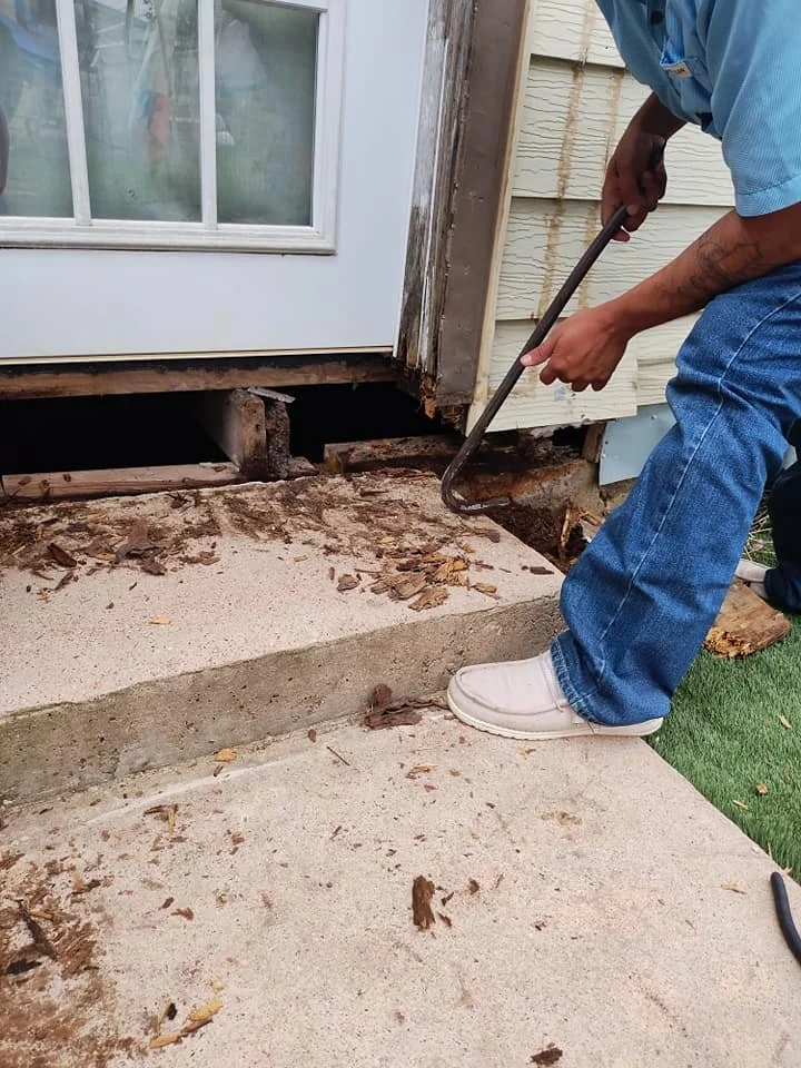 A man wearing blue jeans, a blue shirt, and white shoes is using a crowbar to remove old wood and debris from under a door threshold outside a house. The house has beige siding, and there is a window with a white frame next to the door. The ground is