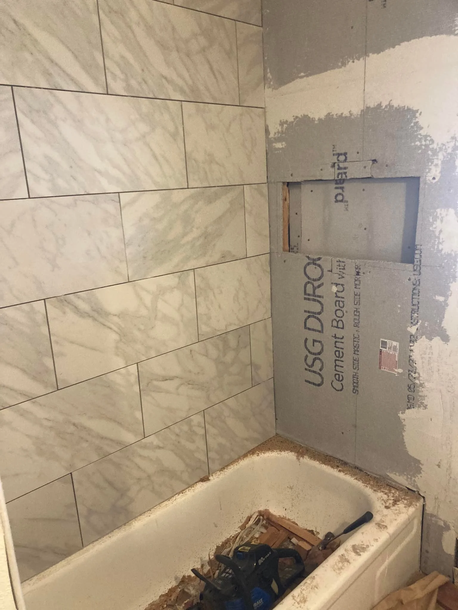 Bathroom under renovation with tiled wall and a bathtub filled with construction tools and debris.