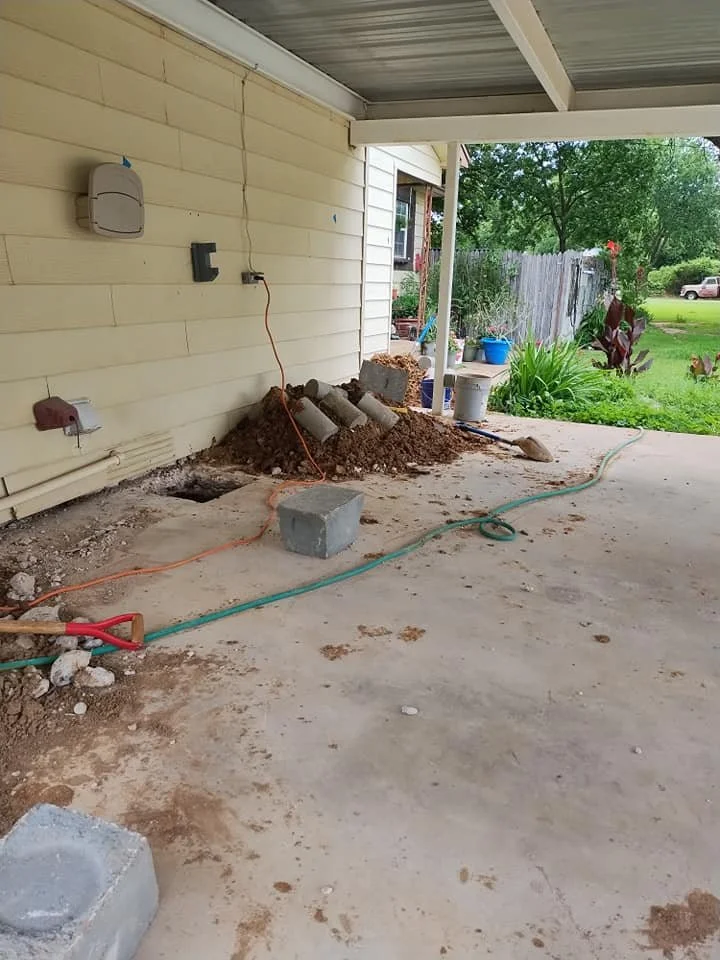 Partially excavated area on a porch, with construction materials and tools, including a pile of dirt, pipes, a concrete block, and garden hoses, with a view of a lawn and trees in the background.