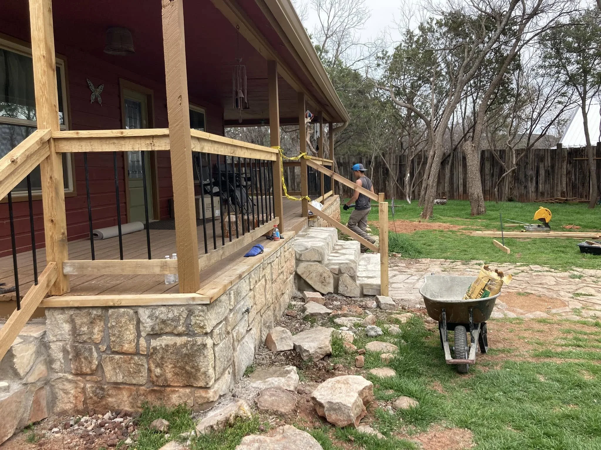 A man installing a wooden porch railing on a stone foundation of a house, outdoor scene with trees and a fence, construction tools and materials scattered around.