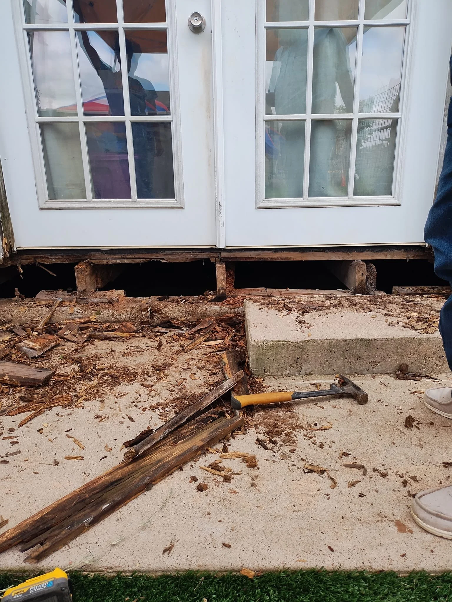 Photo of a house's back door with double glass panel doors, construction work in progress with debris, wood, and tools on the ground, and a concrete step. Part of a person's leg and foot is visible on the right side.