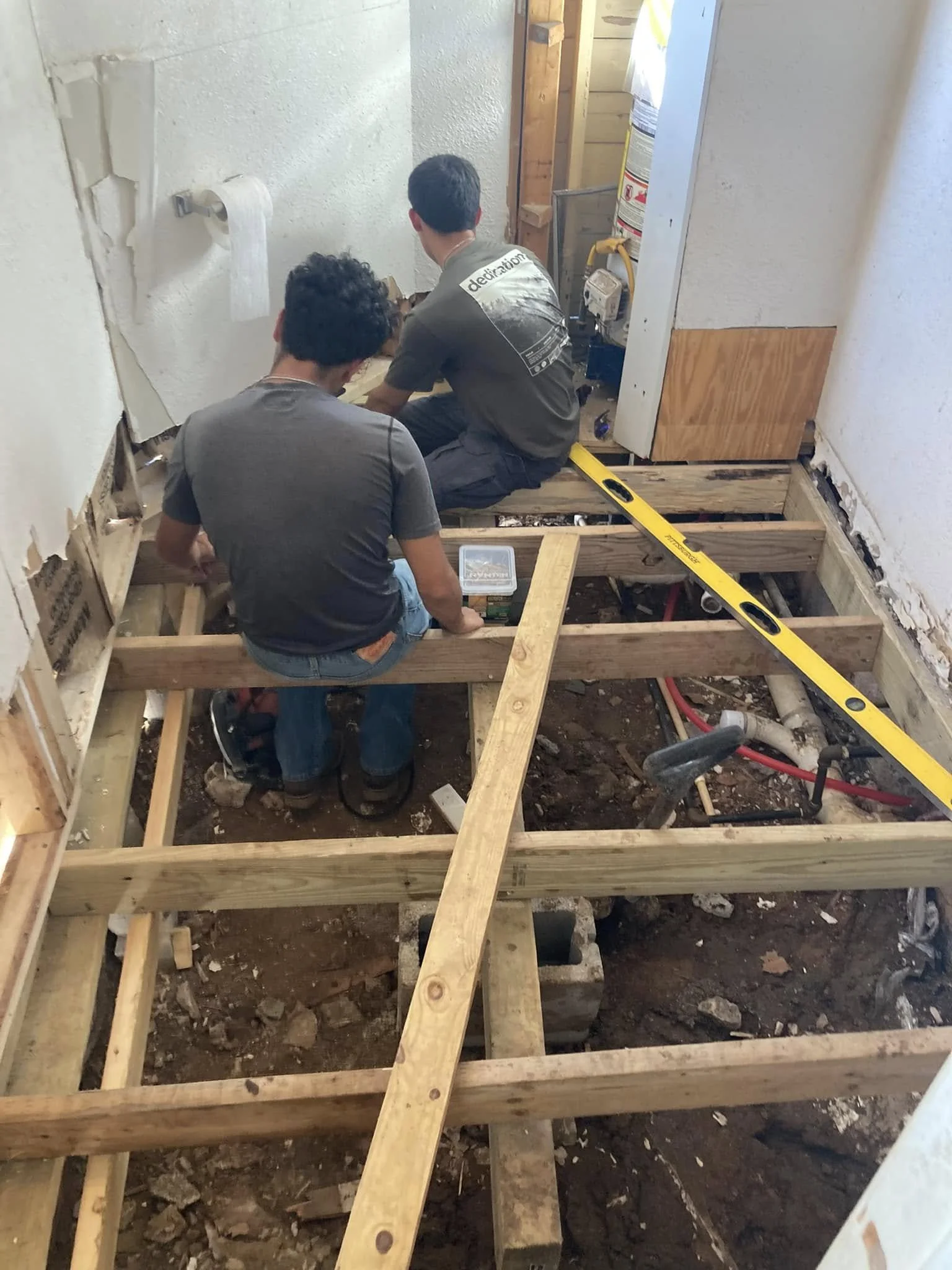 Two men working on a home renovation, installing floor joists in a room with exposed pipes and part of the wall removed.