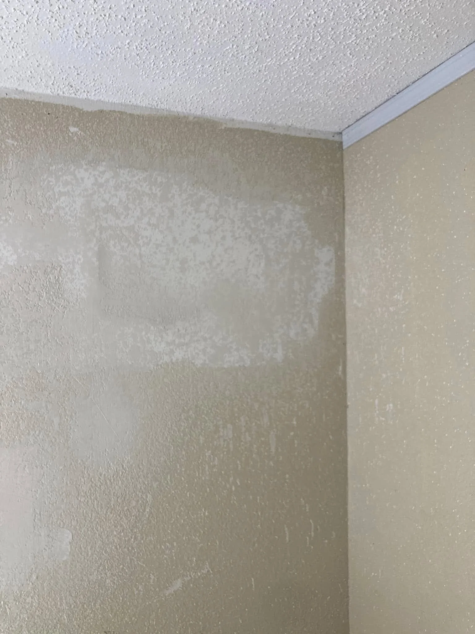 Close-up of a corner of a room with textured beige walls and white ceiling, showing areas of patchy drywall repair.