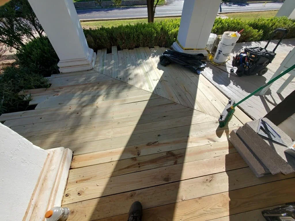A porch undergoing renovation with new wooden flooring installed. Tools, construction materials, and supplies like buckets, a toolbox, a broom, and concrete blocks are visible. Sunlight casts shadows across the area.