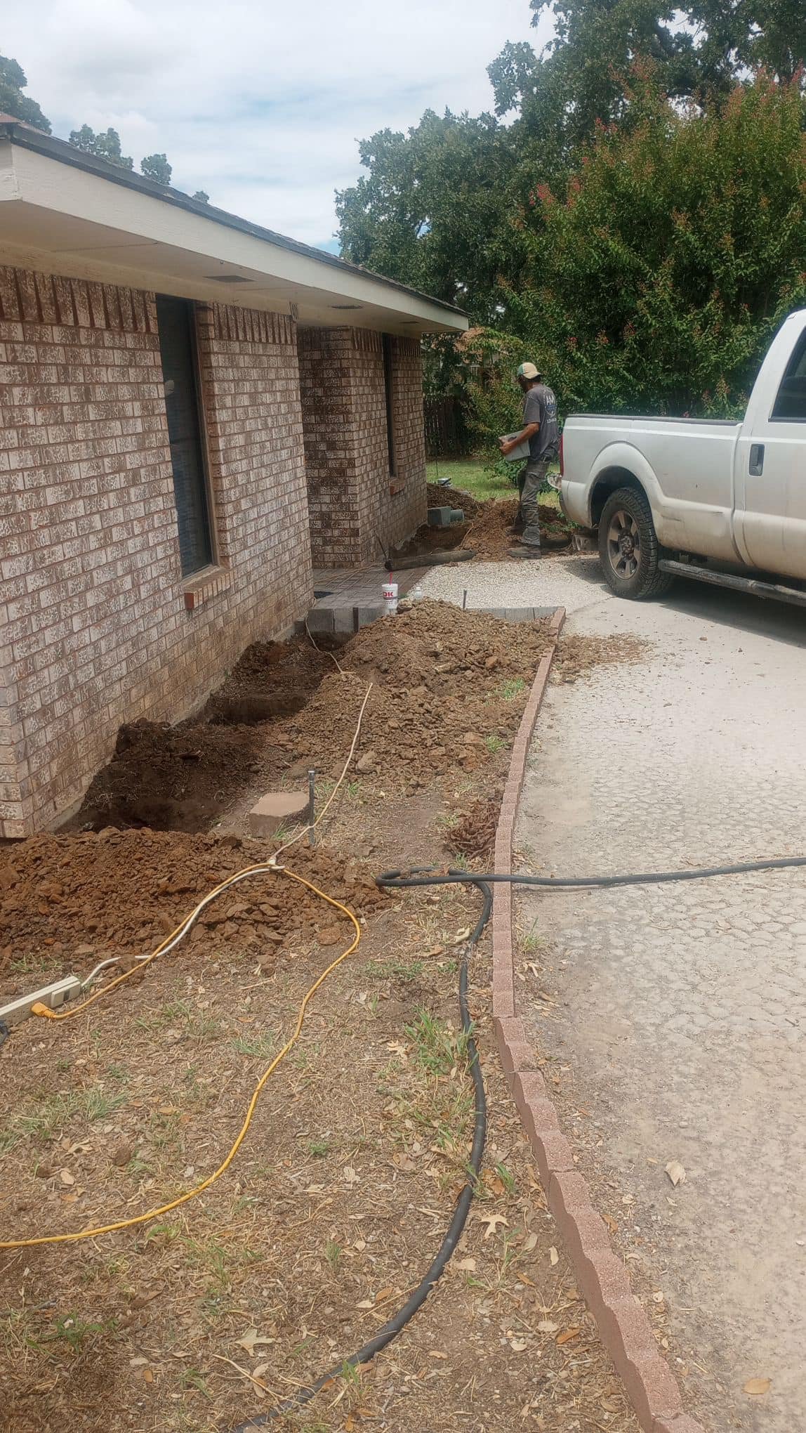 A construction worker is working on the landscaping near a brick house, with a white truck parked on the gravel driveway. The worker appears to be laying bricks or pavers along the edge of the house, with dirt and construction materials surrounding the area.